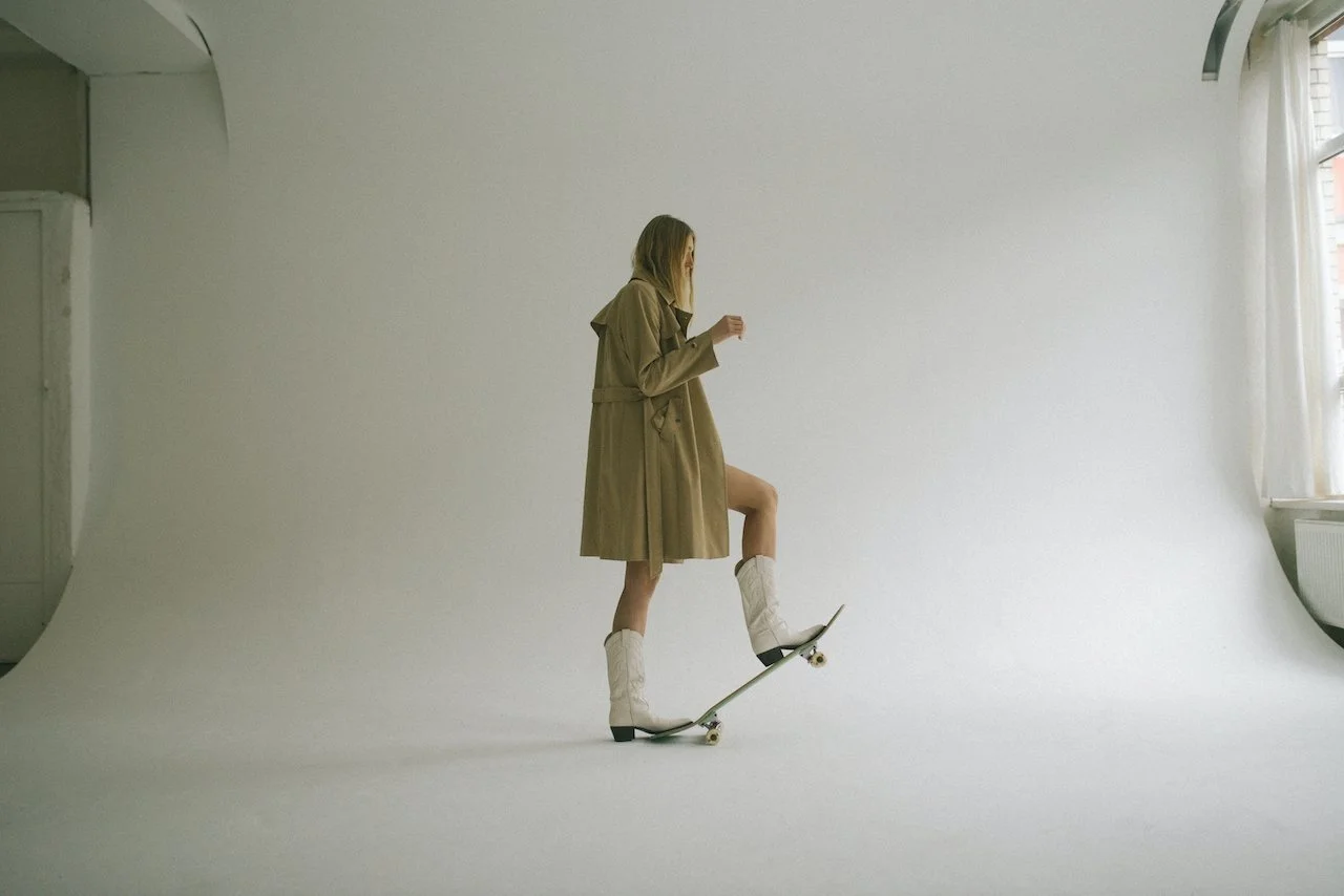 Young woman skating indoors on a skateboard in a minimalist, white studio with natural light from windows.