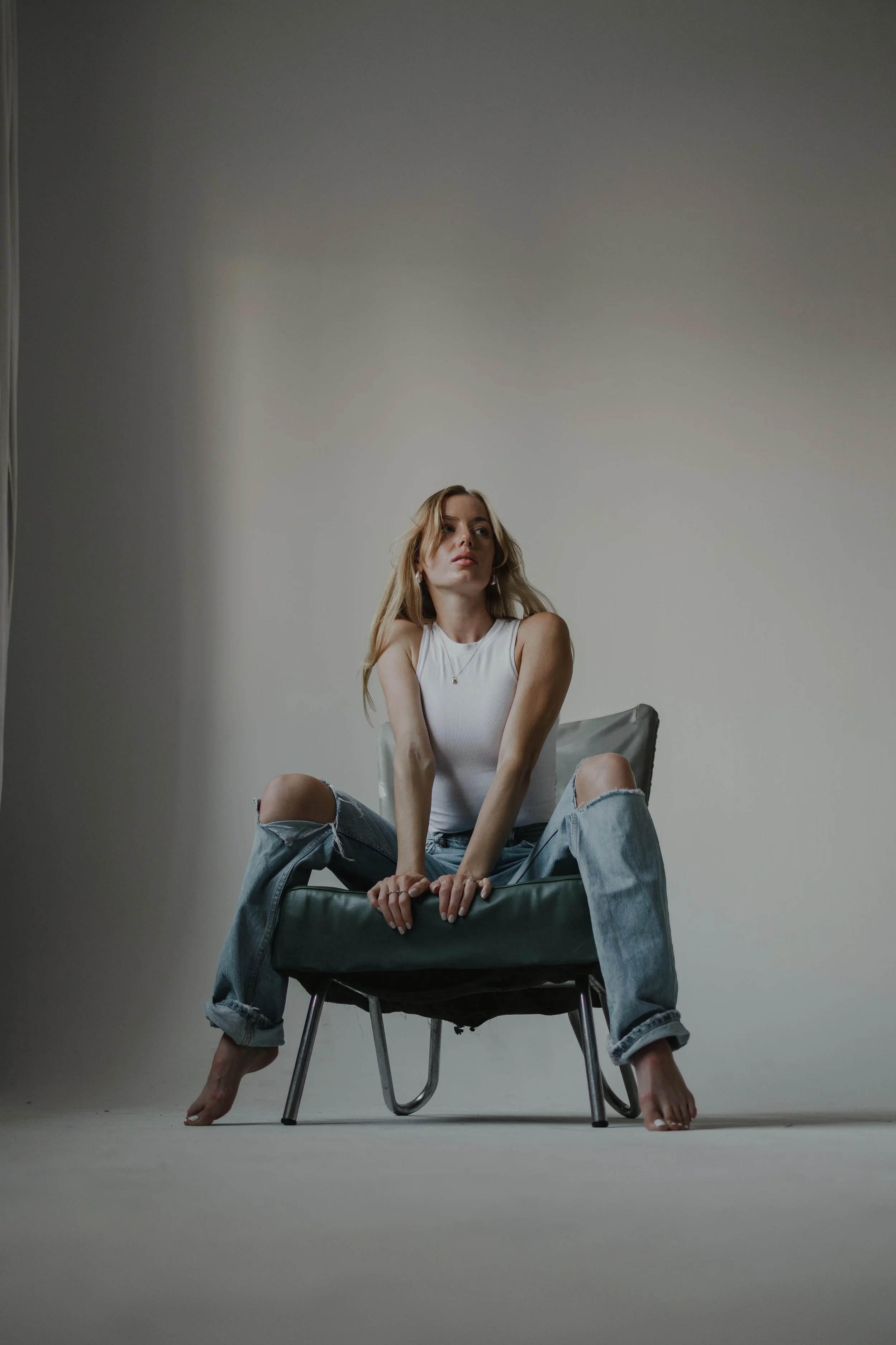 A woman sitting on a chair with her legs spread, wearing ripped jeans and a white tank top, in a minimalistic studio setting.