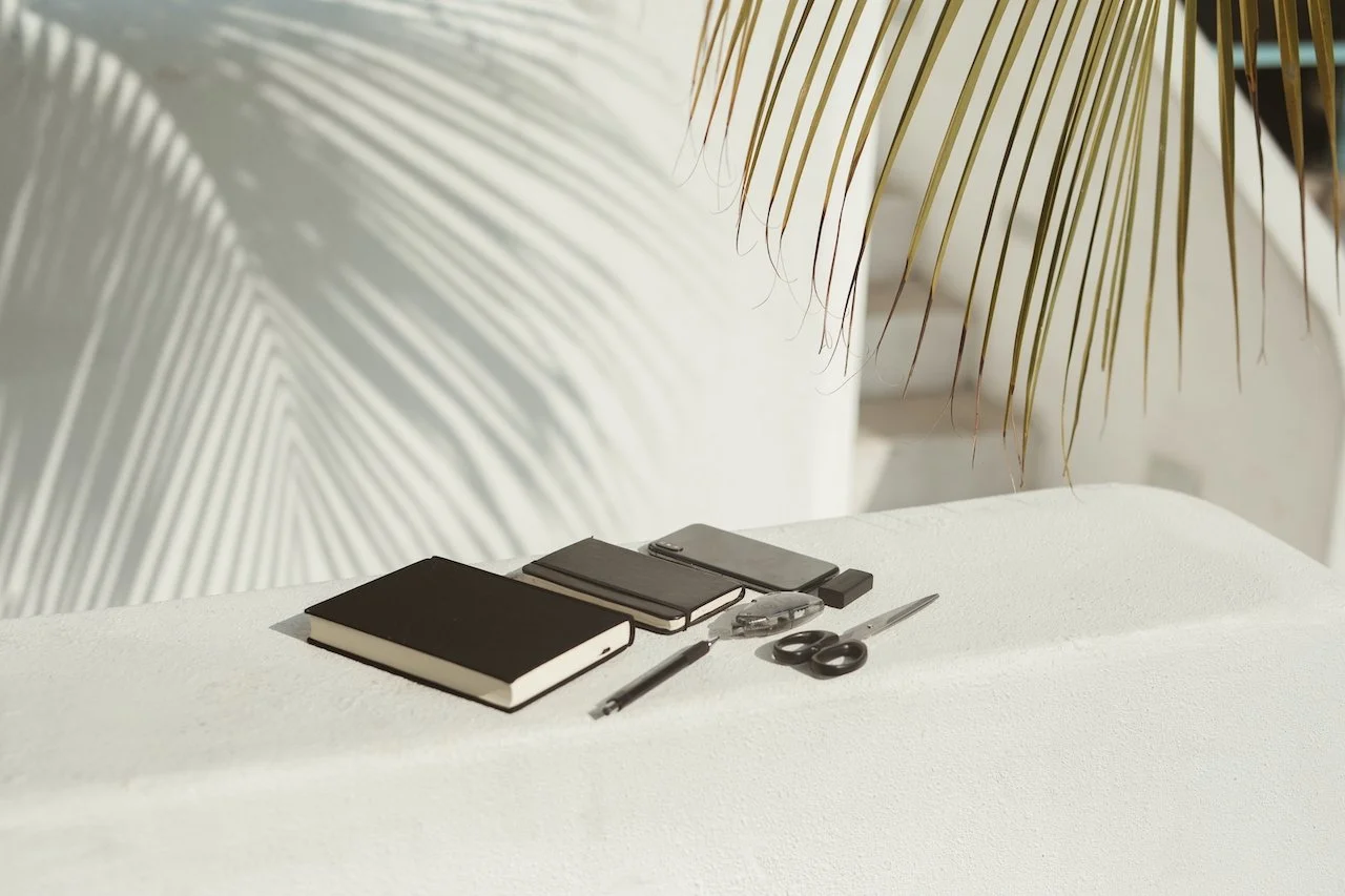 A white table with various office supplies including two notebooks, a smartphone, a USB flash drive, scissors, a pen, and a Swiss Army knife. Palm tree shadows are cast on a white wall in the background.