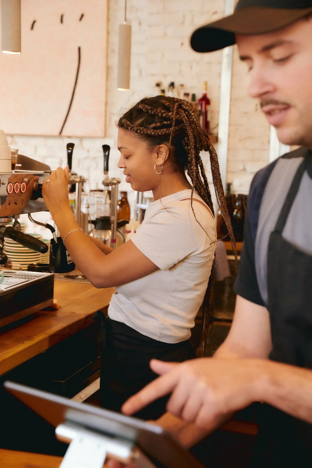A female barista with braided hair making coffee behind the counter at a coffee shop, with a male barista using a tablet in the foreground.