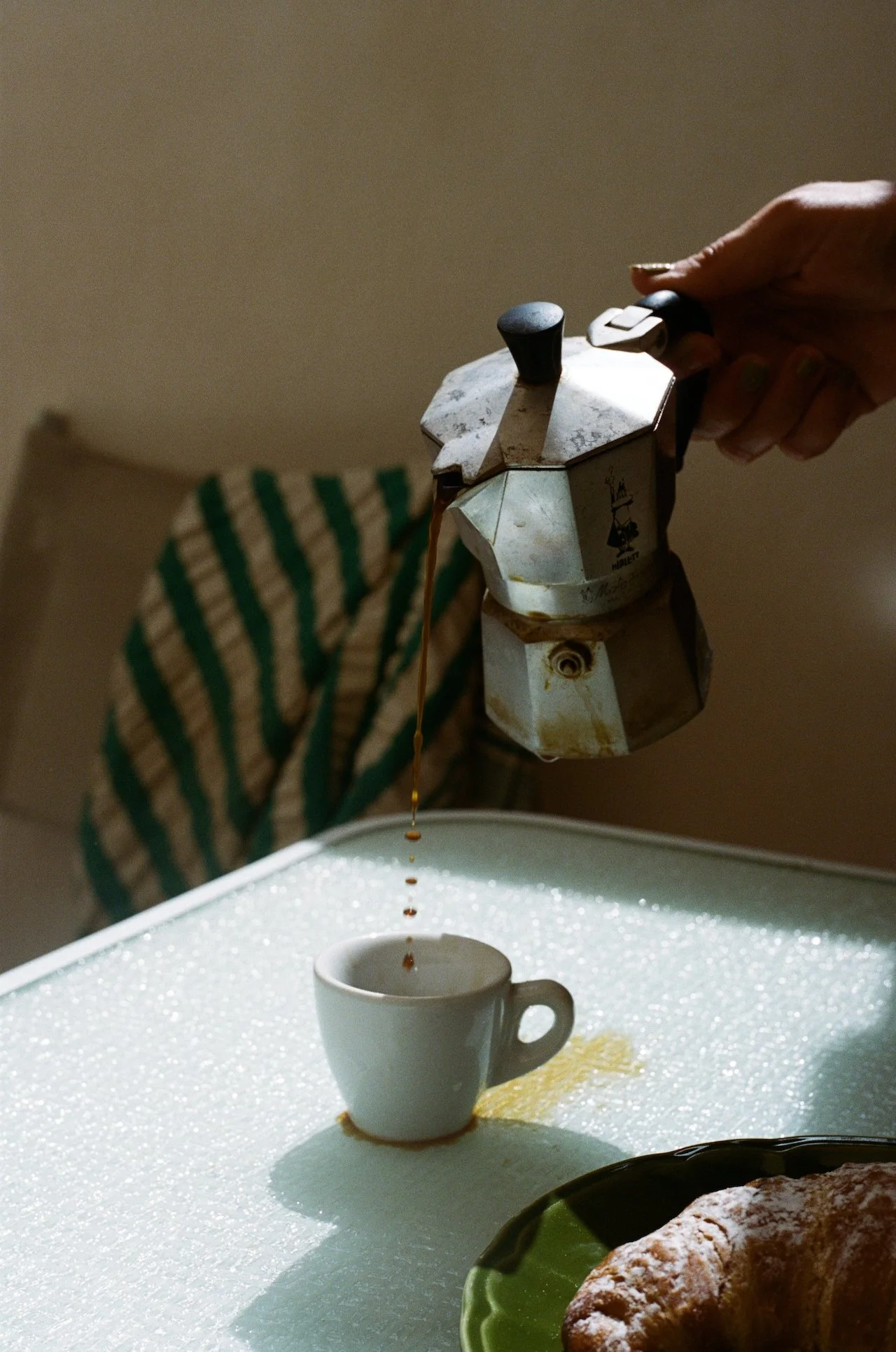 A hand holding a stovetop espresso maker pouring coffee into a small white cup on a textured white tray, with a croissant on a green and black plate nearby.
