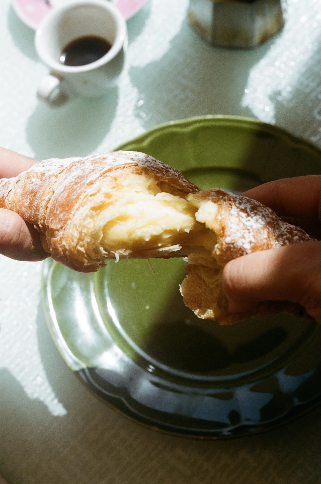 A person holds a croissant filled with cream or custard over a green plate, with a cup of coffee in the background.