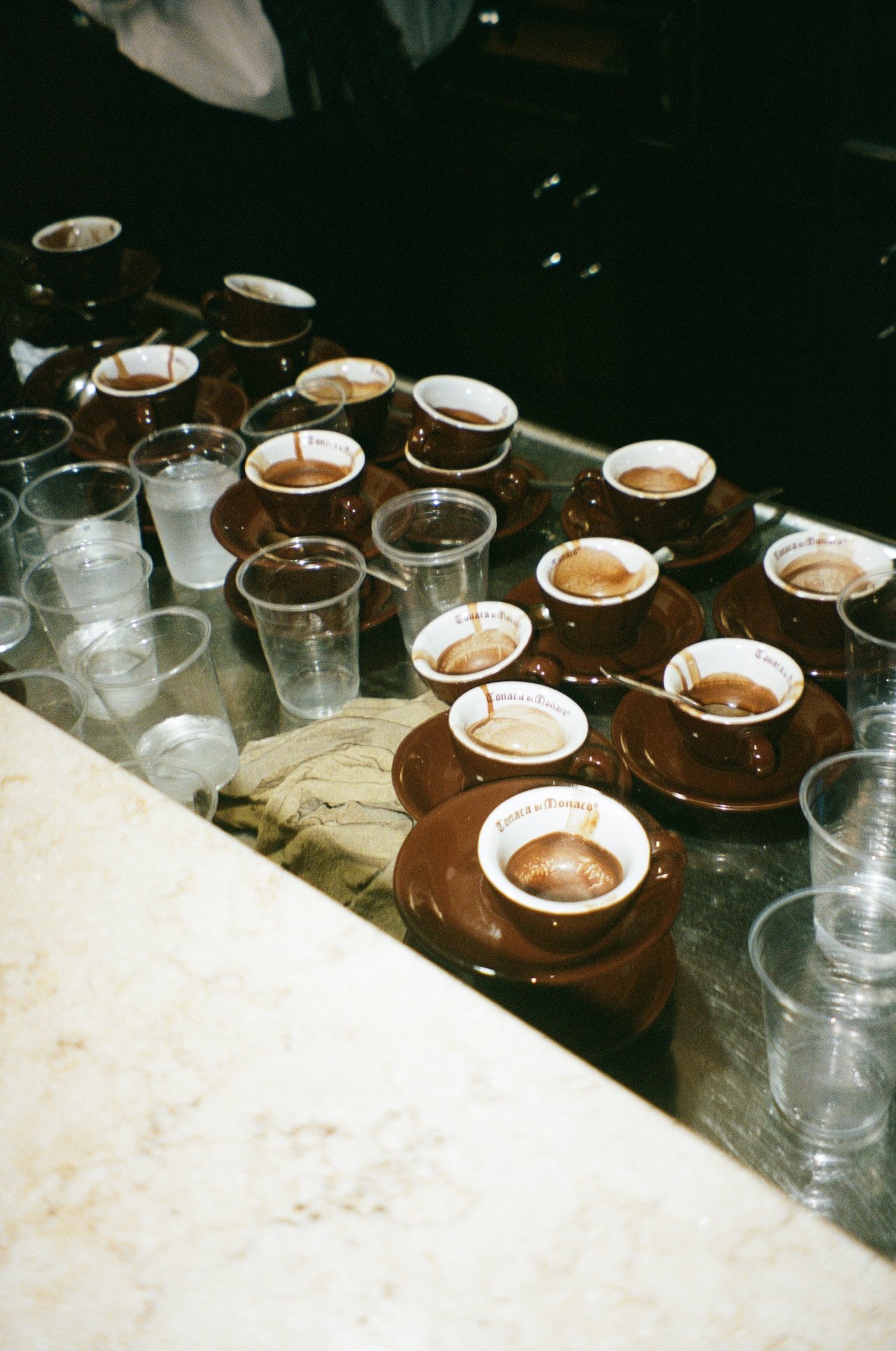 Multiple brown coffee cups with saucers and a few clear plastic cups on a table, some cups contain dark coffee.
