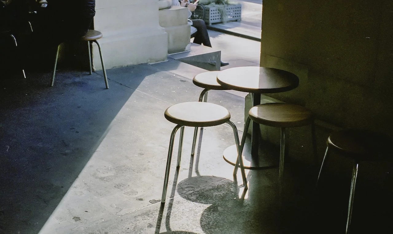 Empty wooden tables and stools in a cafe with sunlight casting shadows on the concrete floor.