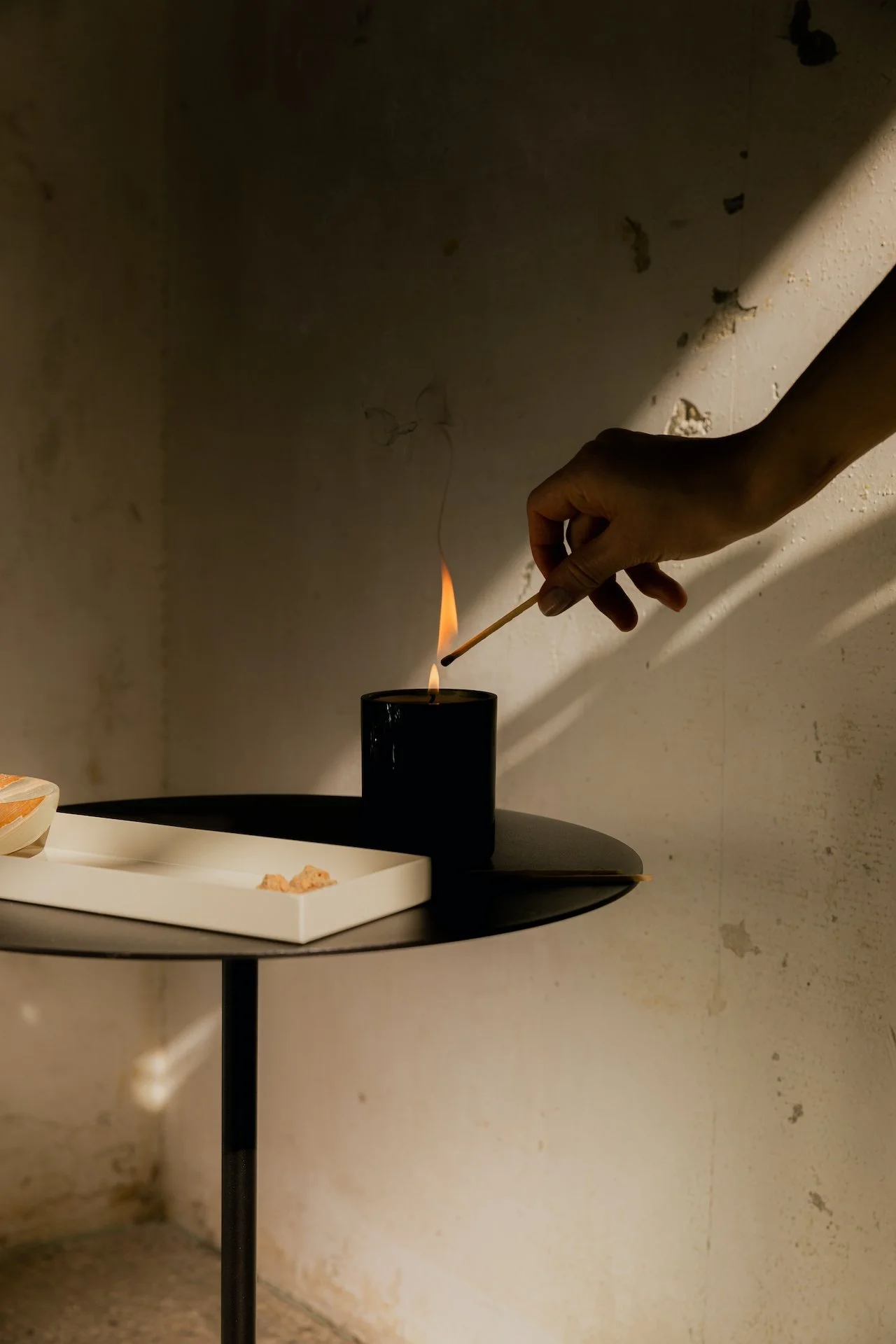 A person's hand is lighting a black candle with a matchstick, on a round black table with a white tray and some snacks, against a plain, textured wall.