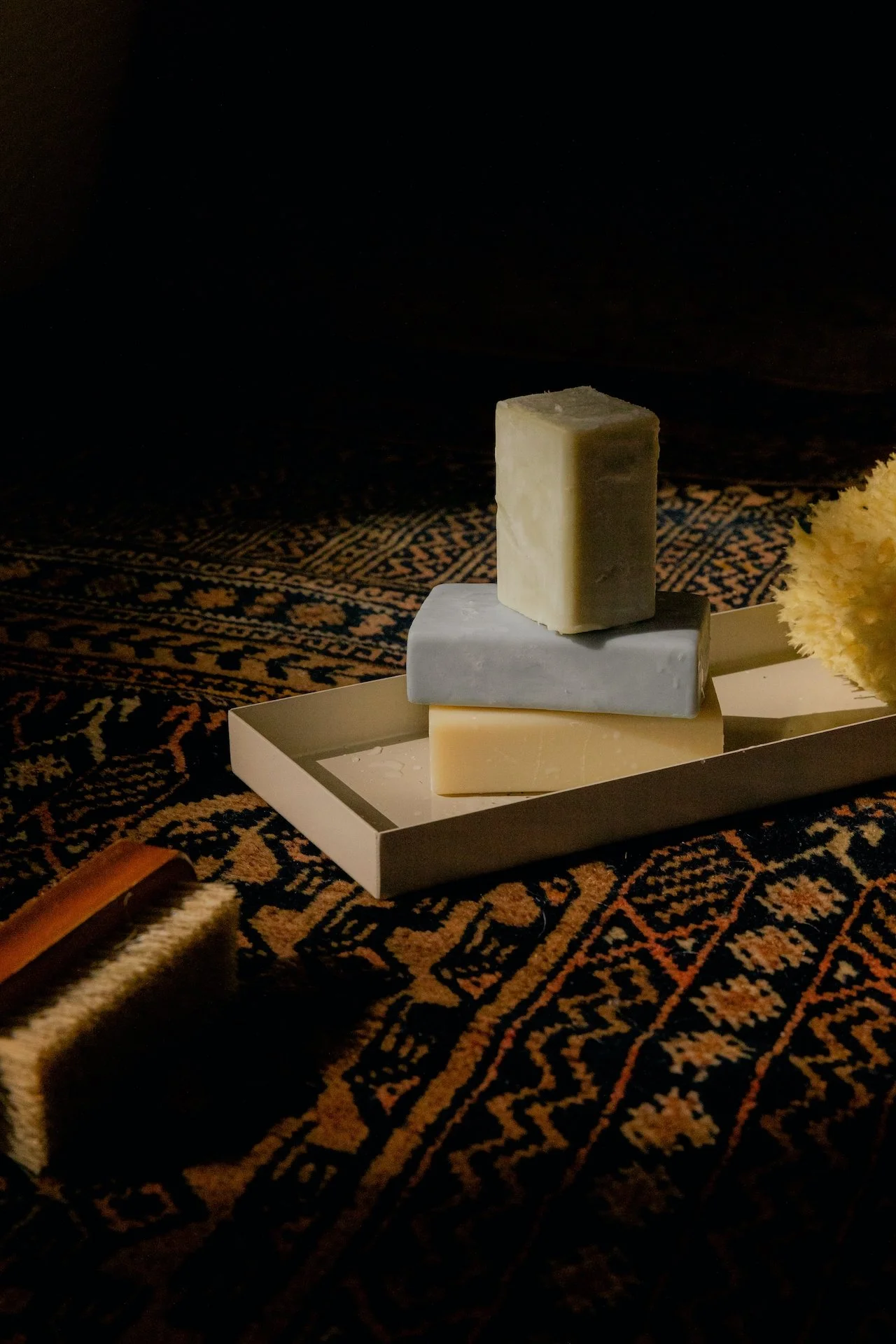 Stacks of three different colored cheeses on a white tray, placed on a decorative dark-patterned surface, with a cheese brush and a yellow flower nearby.