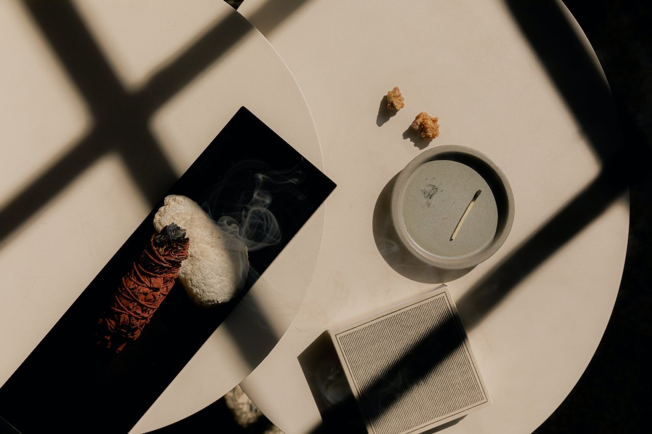 A round white table with a black rectangular tray holding an incense cone, a white stone, and a black dish with smoke. Next to the tray, there's a small beige candle, two small rocks, and a closed white book. Shadows from window blinds cast lines acr