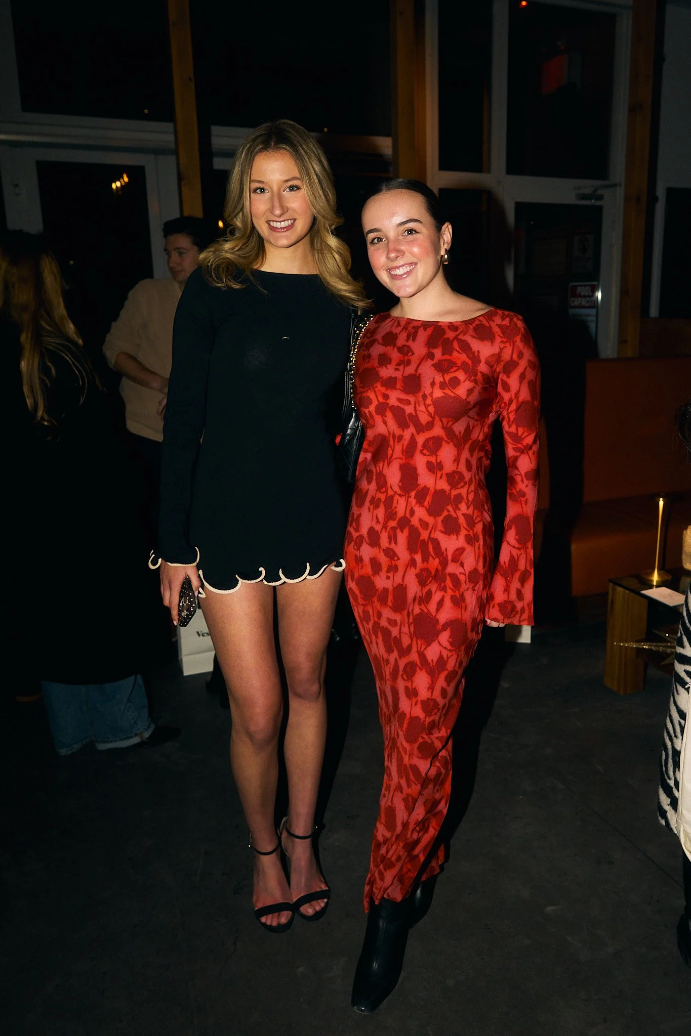Two women smiling and posing together at an indoor event, one in a black dress and the other in a red patterned outfit, with other people in the background.