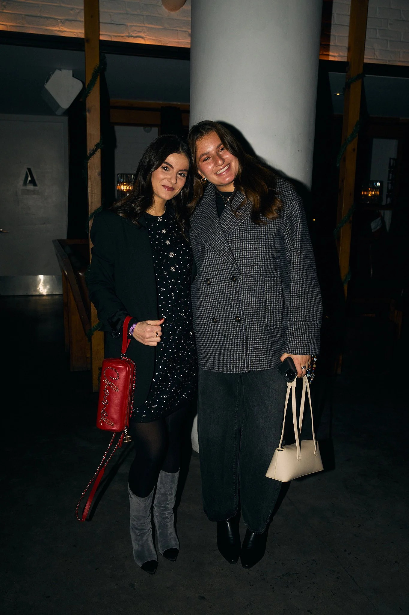 Two women standing indoors near a white pillar, smiling for the camera. One woman has dark hair and wears a black dress with a black blazer and gray suede boots, holding a red purse. The other woman has brown hair and wears a gray checkered coat with