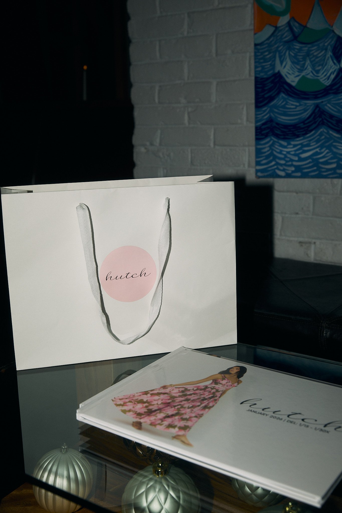 A white shopping bag with pink circle and the word 'hutch' on it, a photo album with a woman in a floral pink dress, and decorative ornaments on a glass table.