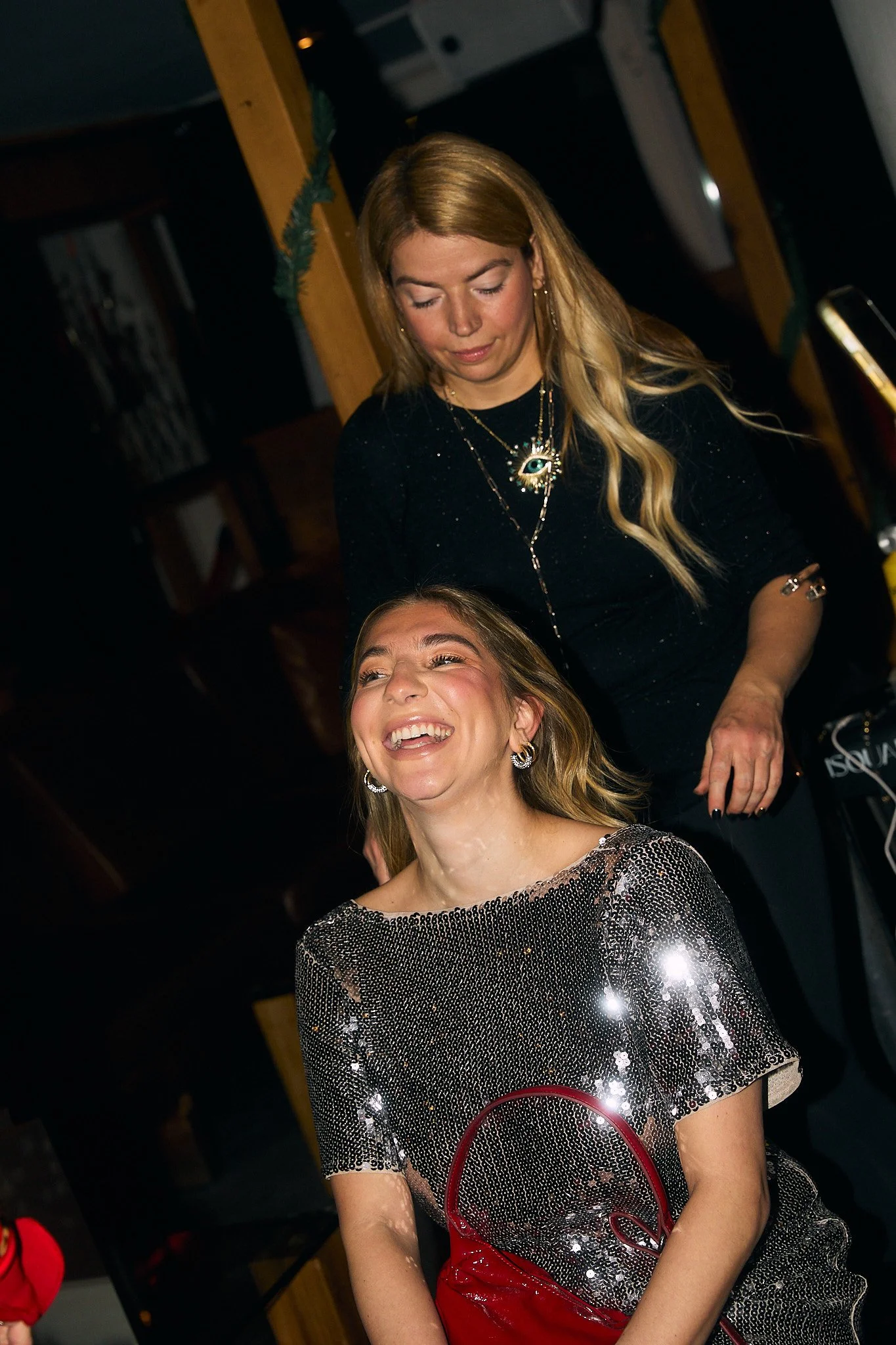 Two women, one seated and one standing behind her, at a festive event. The seated woman is smiling and wearing a sparkly dress with earrings and a red handbag. The standing woman has long blonde hair, wearing a black top with jewelry, and is looking 