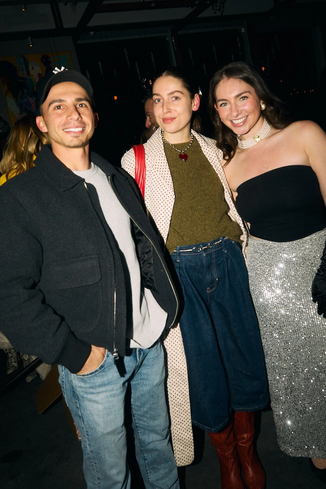 Three people at a social gathering, smiling and posing for the camera. The person on the left is a man wearing a black jacket, white shirt, and baseball cap. The person in the middle is a woman with dark hair, wearing a polka dot blazer, green top, a