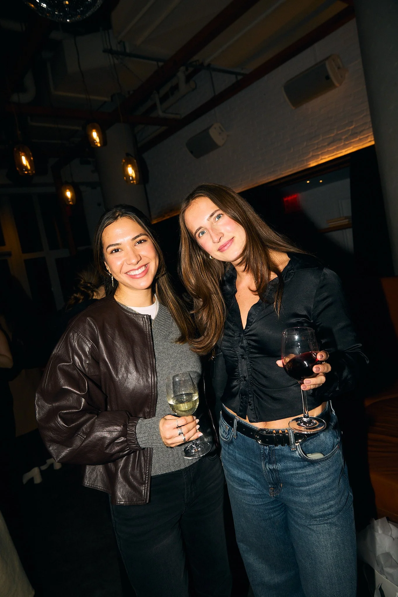 Two women smiling and holding glasses of wine in a dimly lit indoor setting.