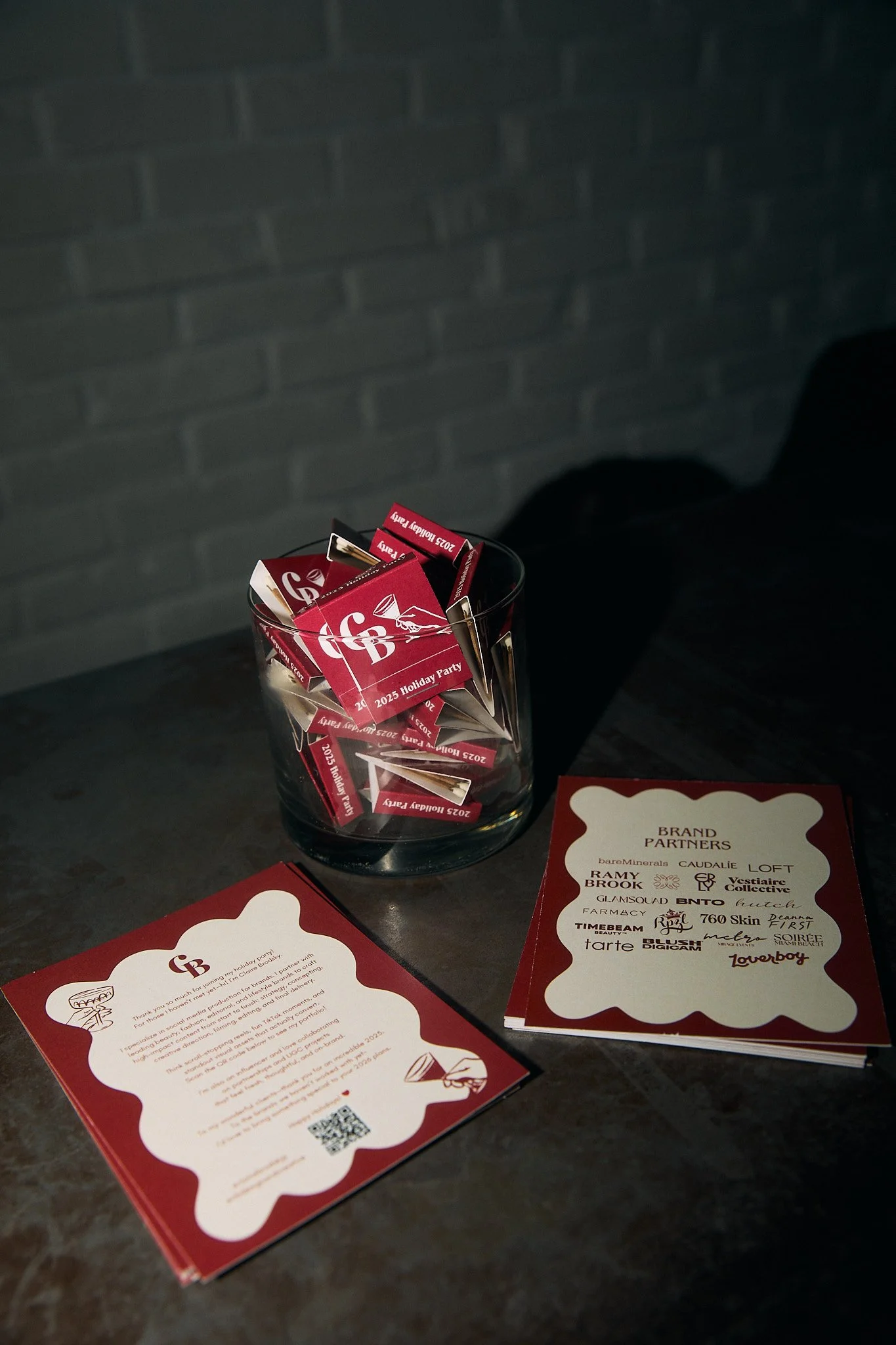 A glass container filled with small red and white cards labeled '2023 Holiday Party,' on a dark surface with a sheet and a card with a list of brand partners next to it.