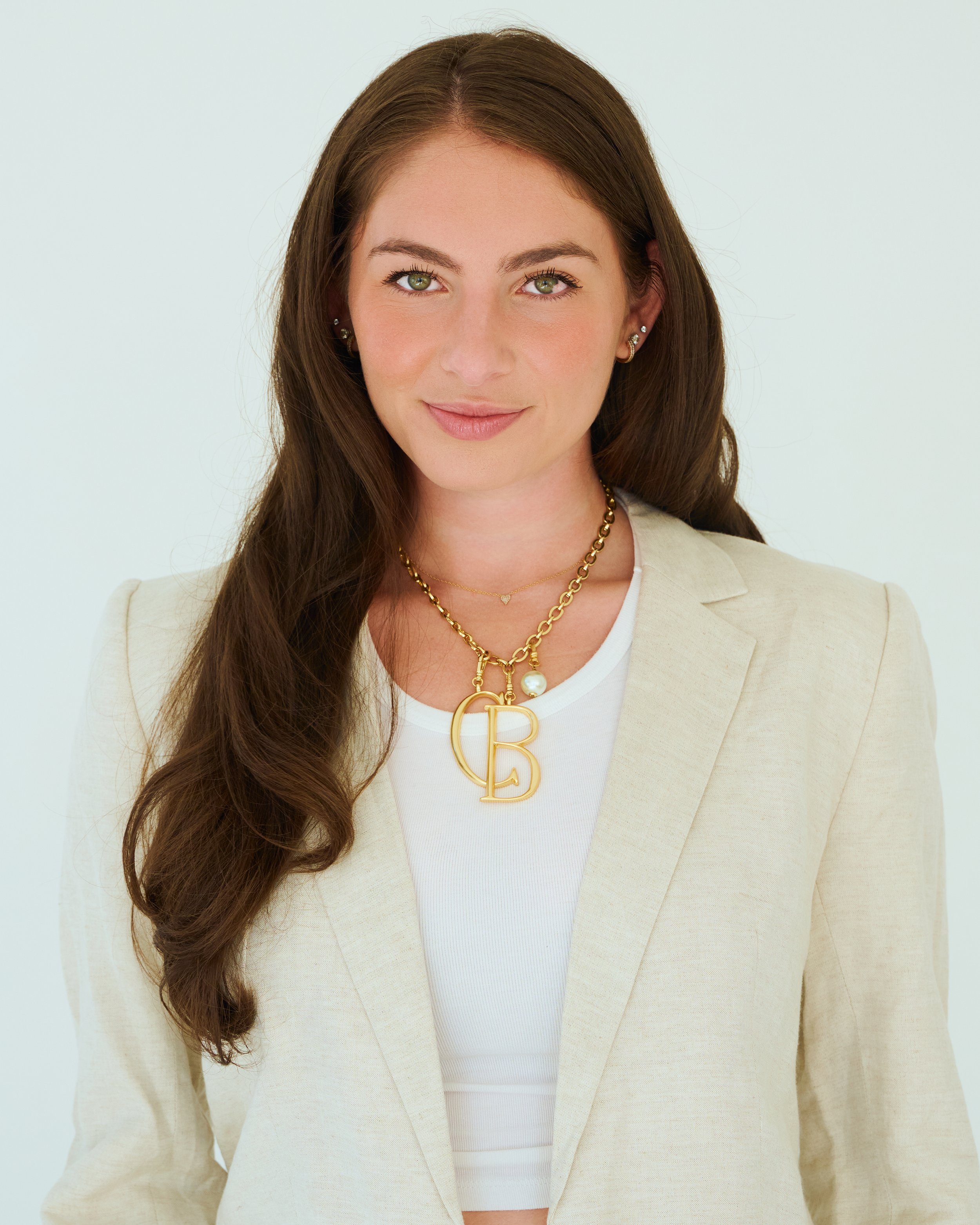 Portrait of a woman with long brown hair, green eyes, wearing a beige blazer, white top, gold jewelry, and smiling at the camera.