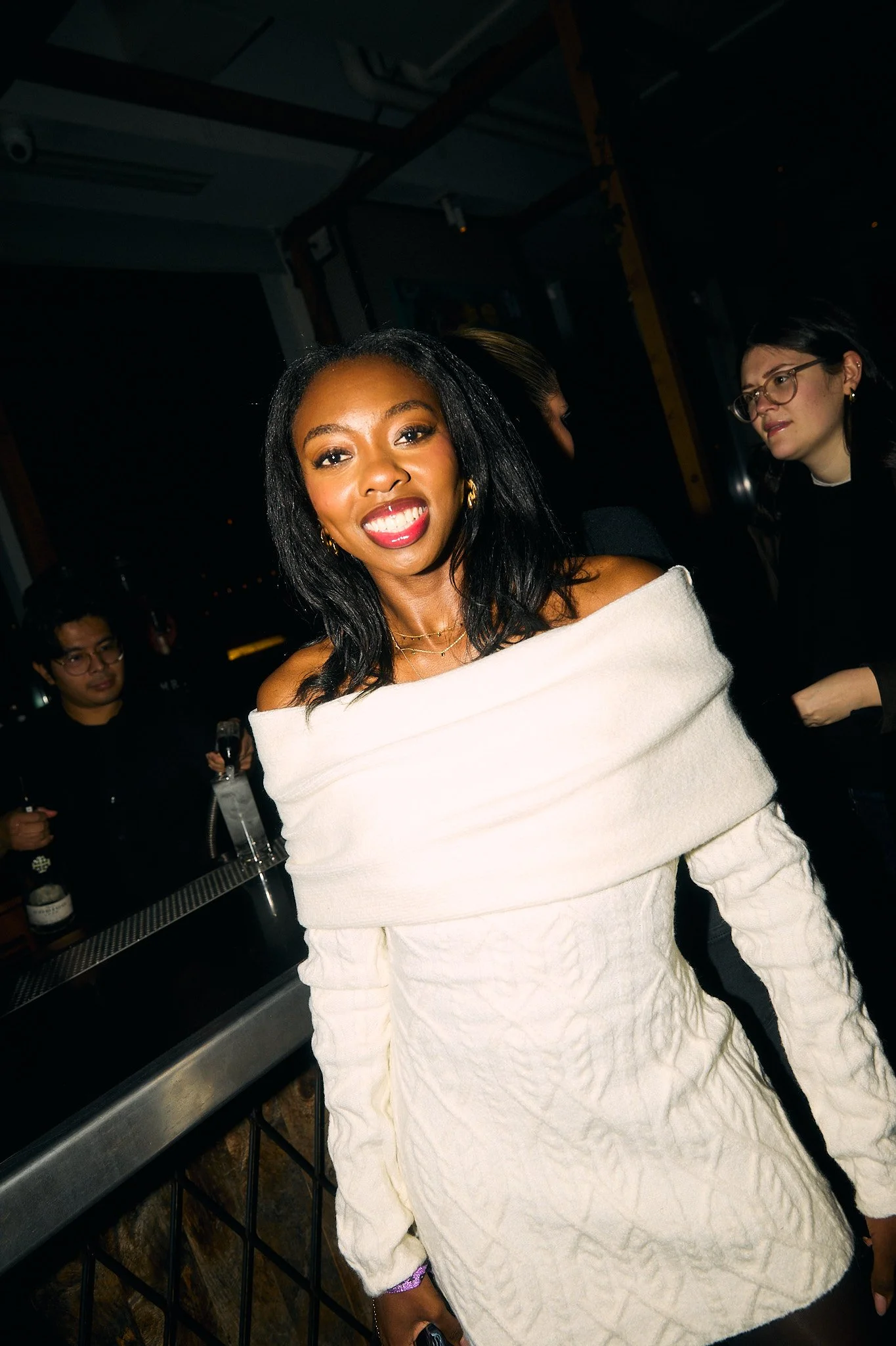 A woman smiling at a social gathering, wearing an off-the-shoulder white dress with textured fabric, with people and a bar in the background.