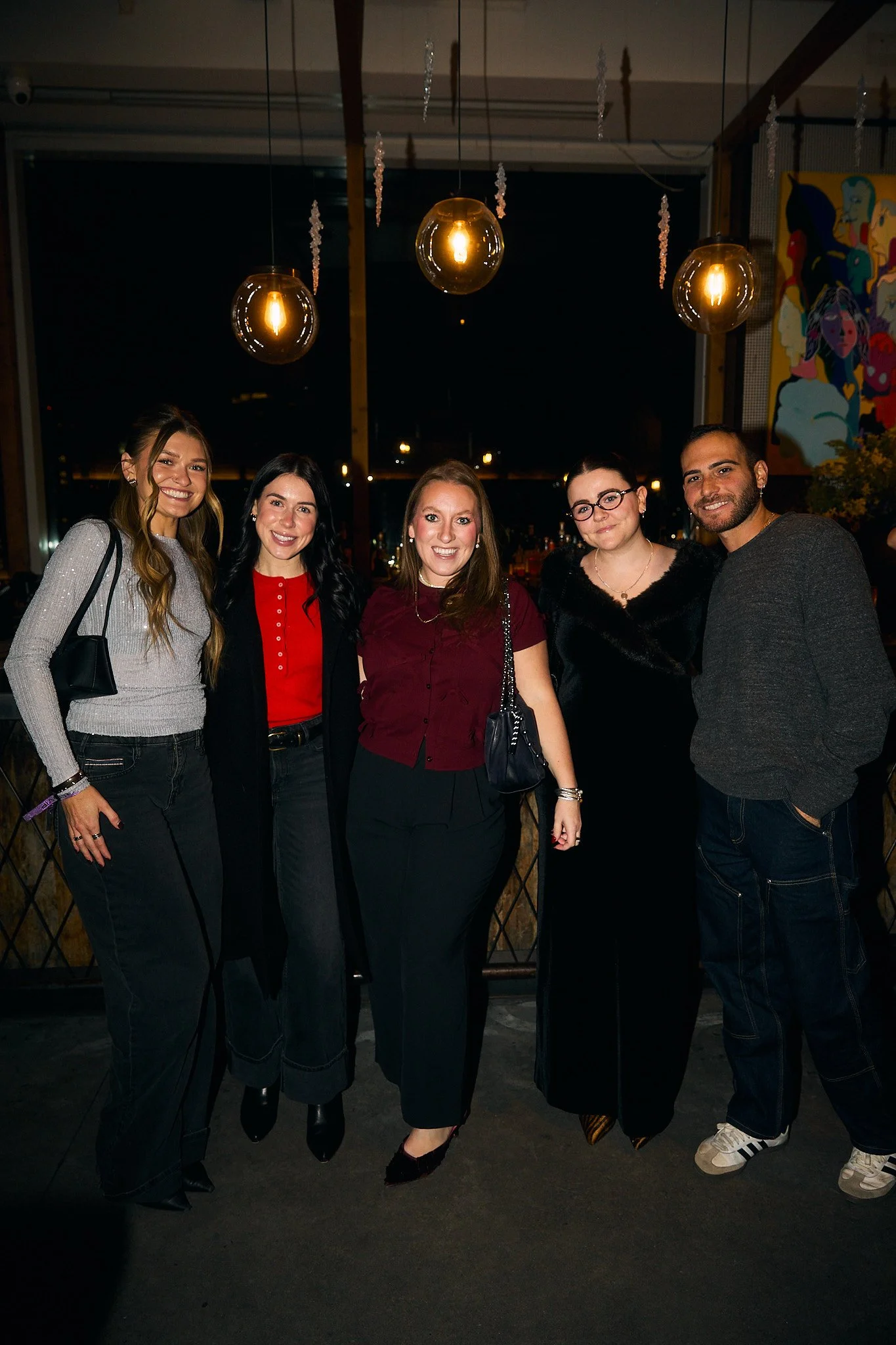Five people standing together in a dimly lit indoor setting at night, smiling for a photo, with decorative hanging lights and wall art in the background.