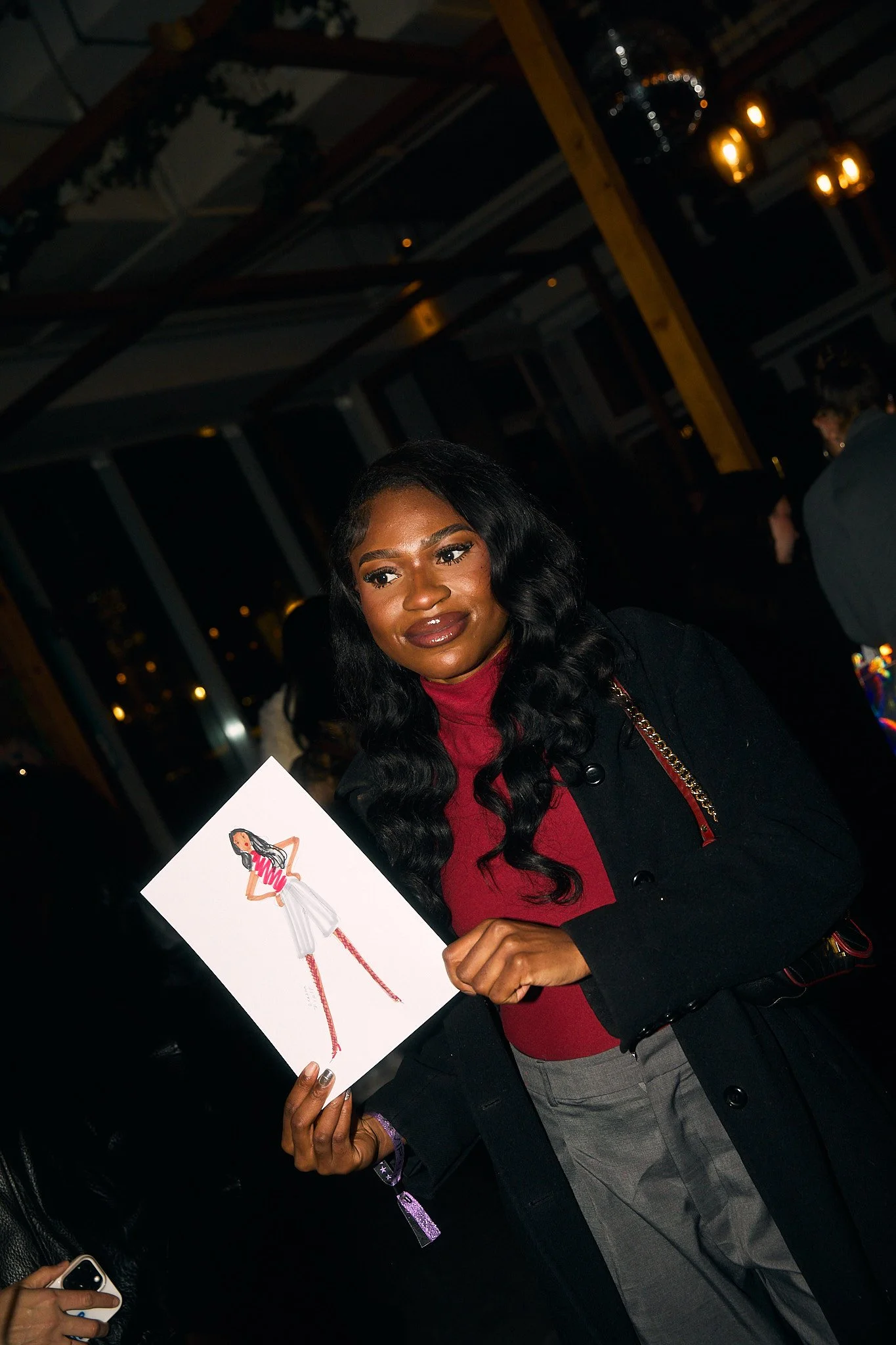 A woman with long black hair, wearing a red turtleneck and black coat, is holding a drawing of a woman in a red and white striped top, white skirt, and red boots. She is smiling and standing indoors at a social event.