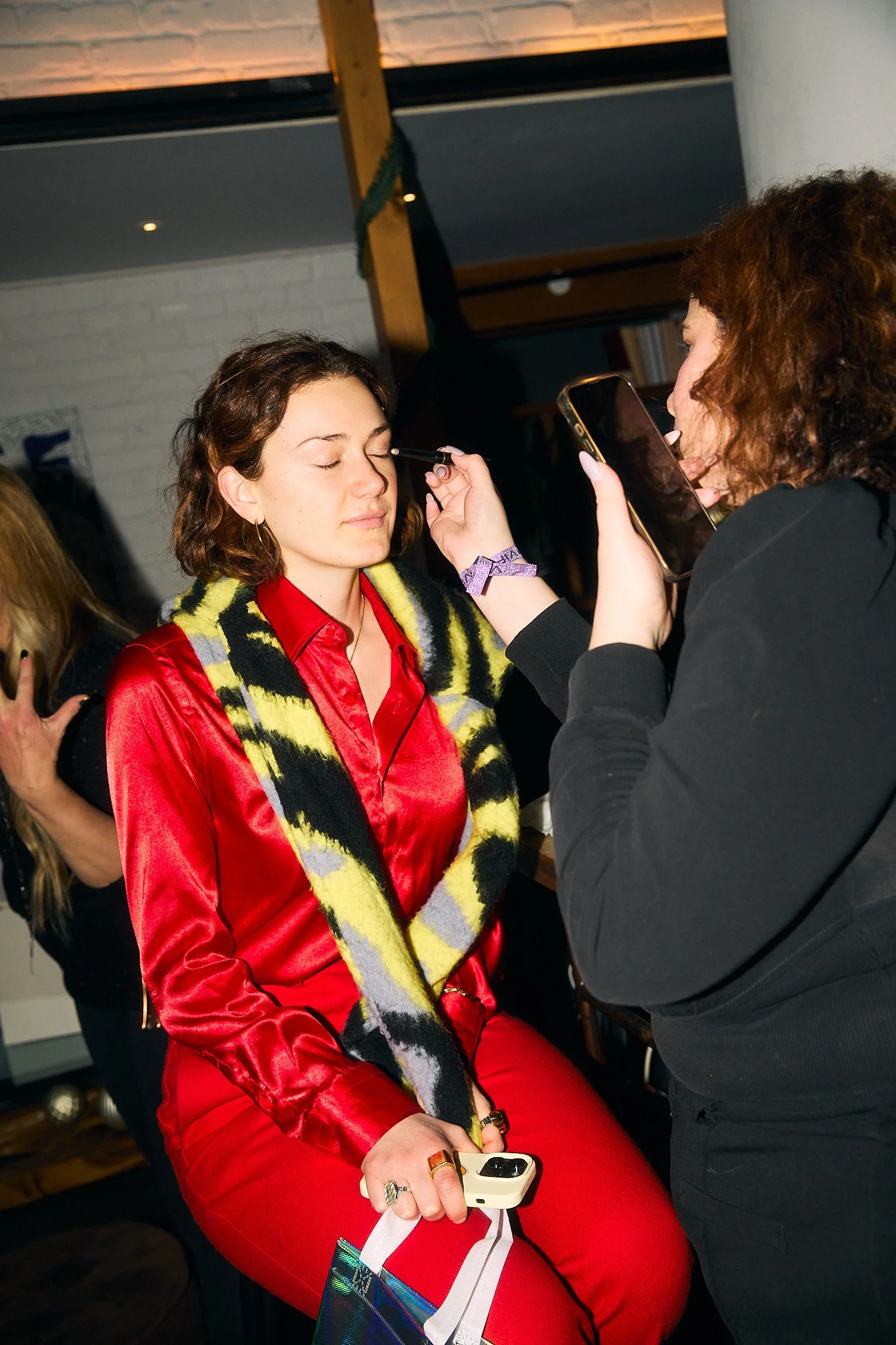A woman in a red satin outfit and a colorful scarf sitting with her eyes closed while a makeup artist applies makeup to her face, holding a makeup brush and a phone.