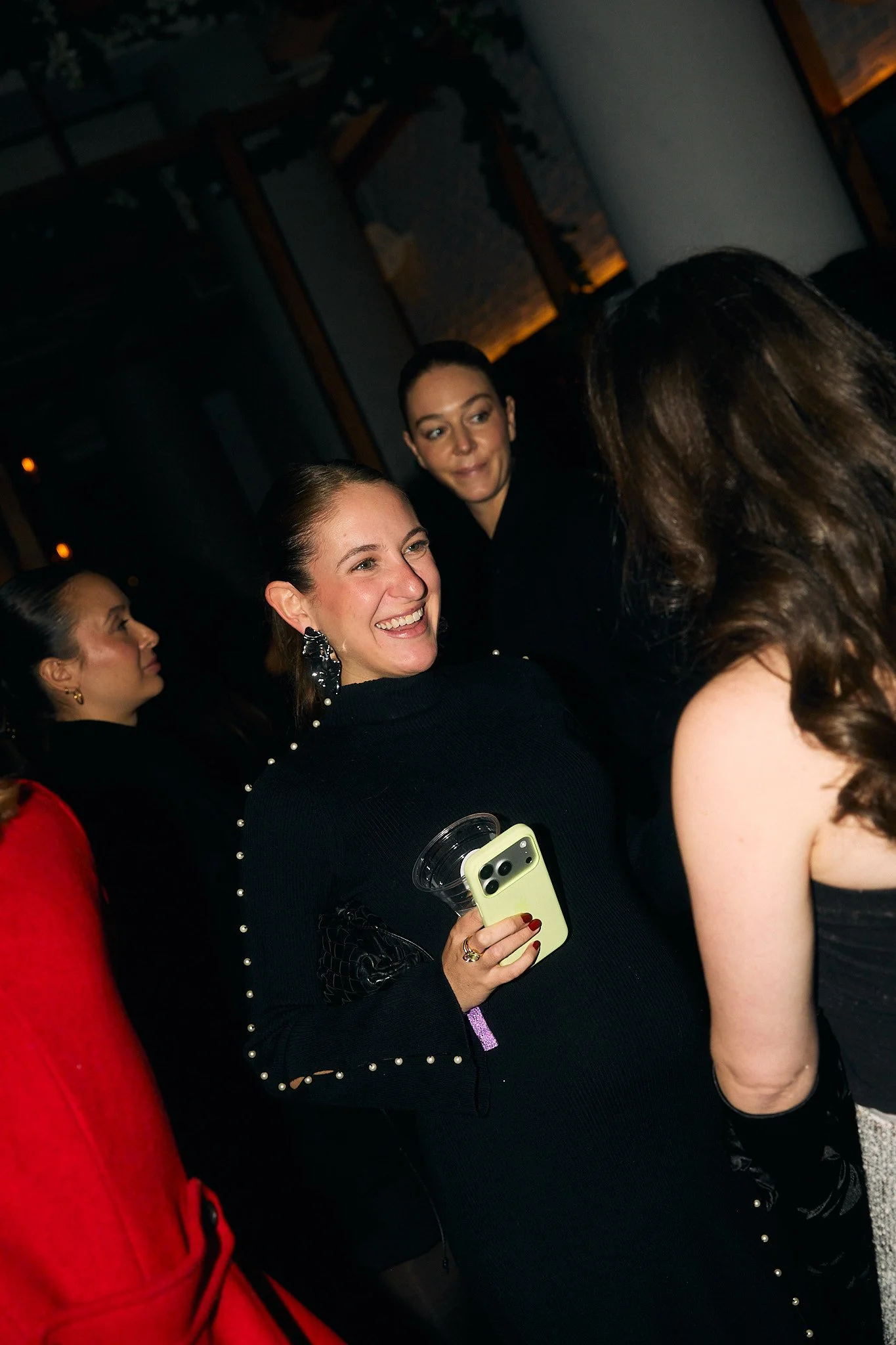 Group of women socializing indoors, one woman in the center smiling, holding a phone and a drink, wearing a black top with pearl embellishments, surrounded by others.