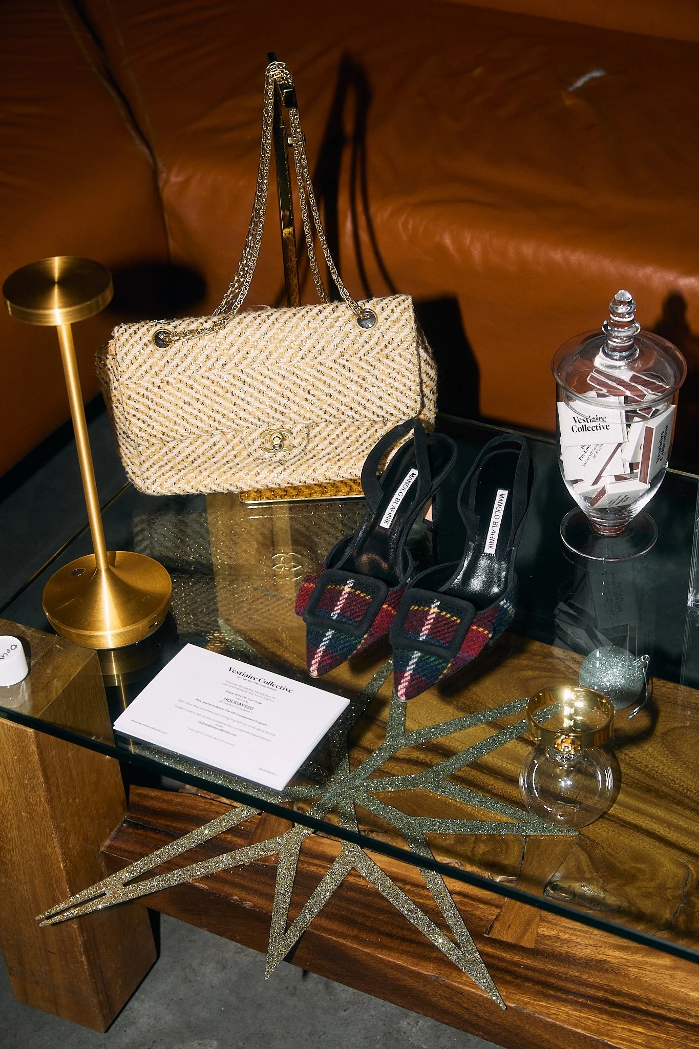 A display table with a beige handbag with chain handles, a pair of plaid high heels, a clear glass jar with a lid, a glittery star-shaped decoration, and a white card, all set against a brown leather sofa background.
