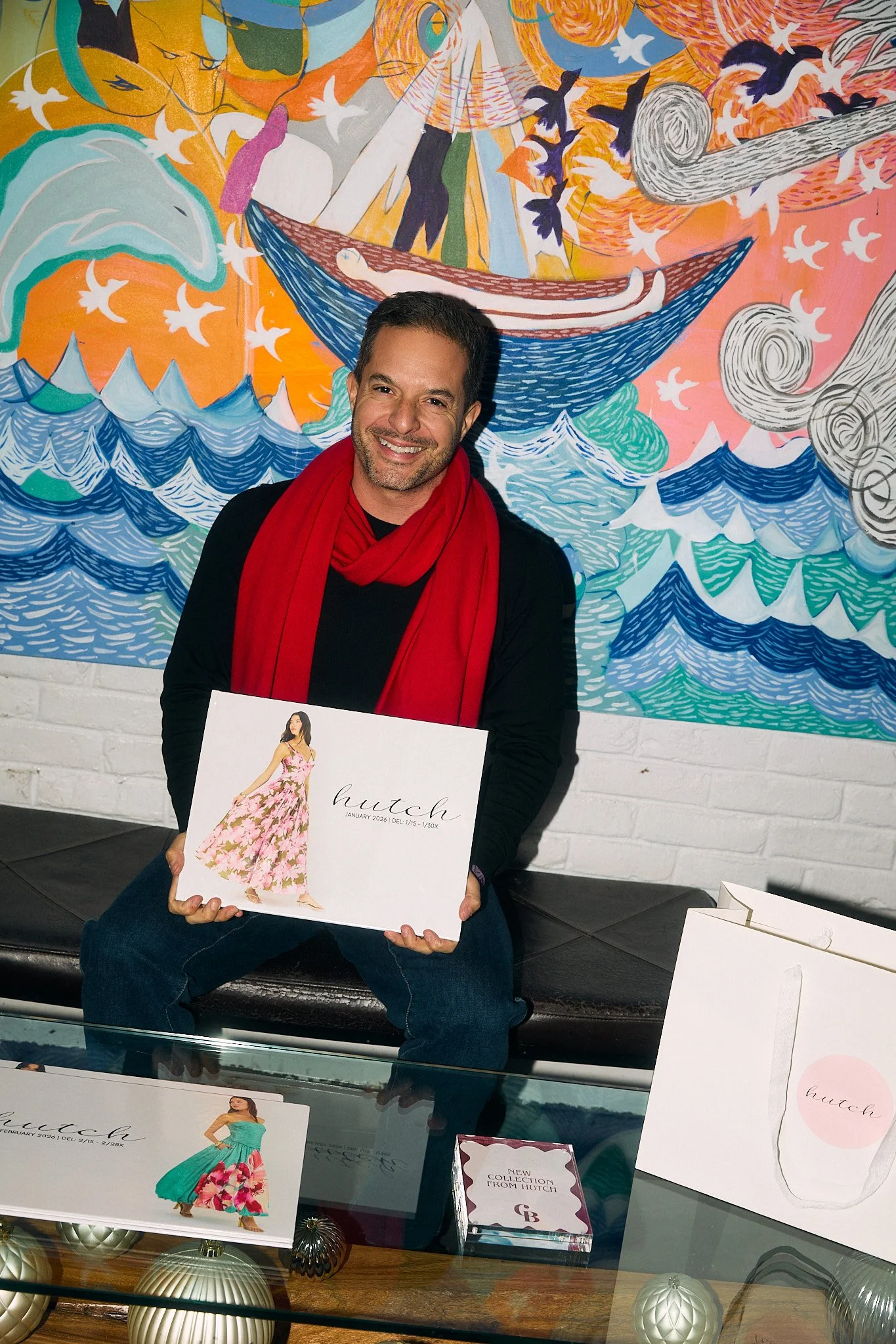 A man with a beard and dark hair, wearing a black shirt and a red scarf, smiling and holding a white box with a pink floral dress on it. He is sitting on a black bench in front of a colorful, abstract mural featuring a boat, waves, birds, and swirls.