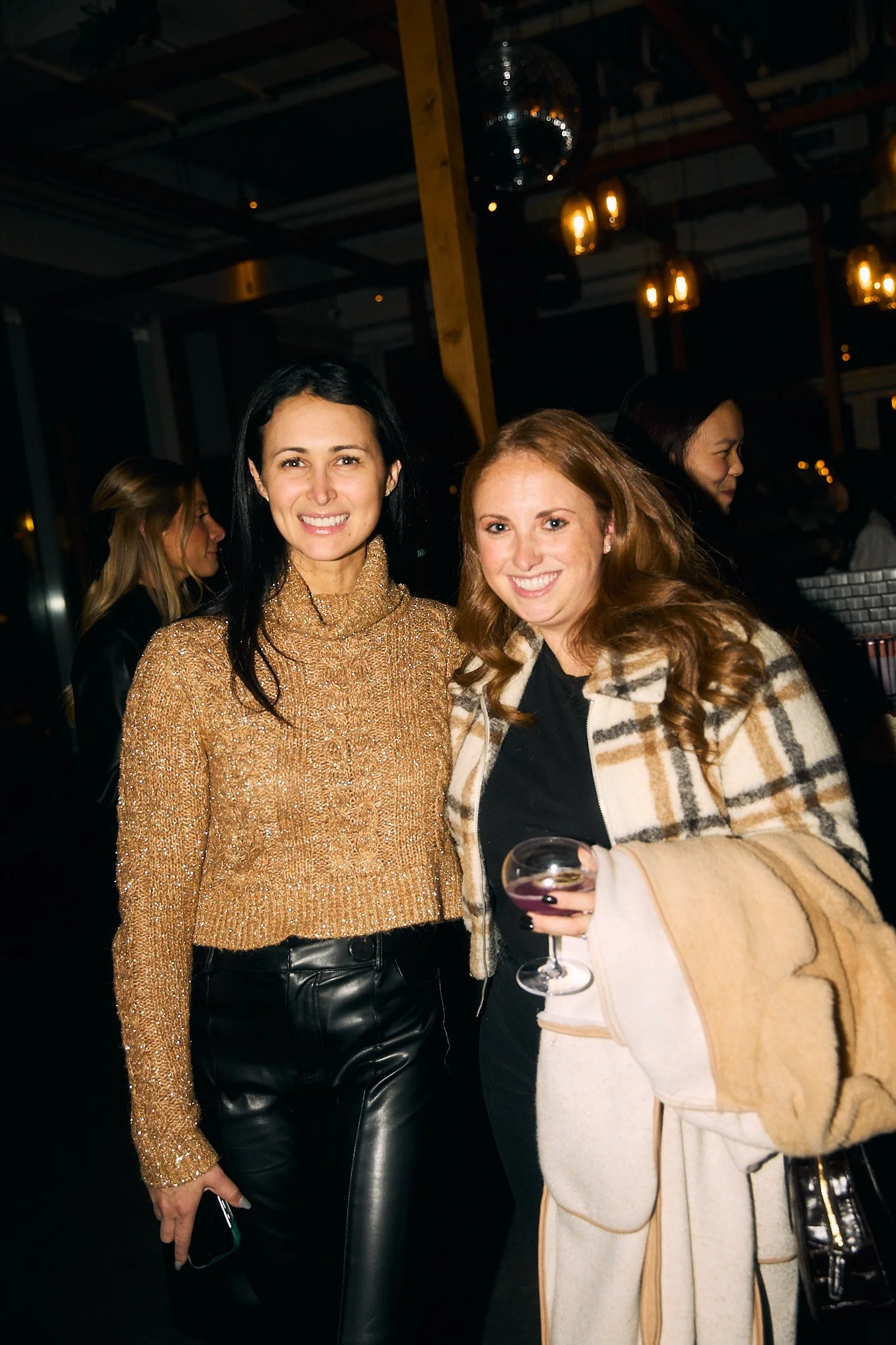 Two women smiling at a social gathering inside a dimly lit venue, one holding a glass of wine and the other with a smartphone in hand, wearing stylish clothing.