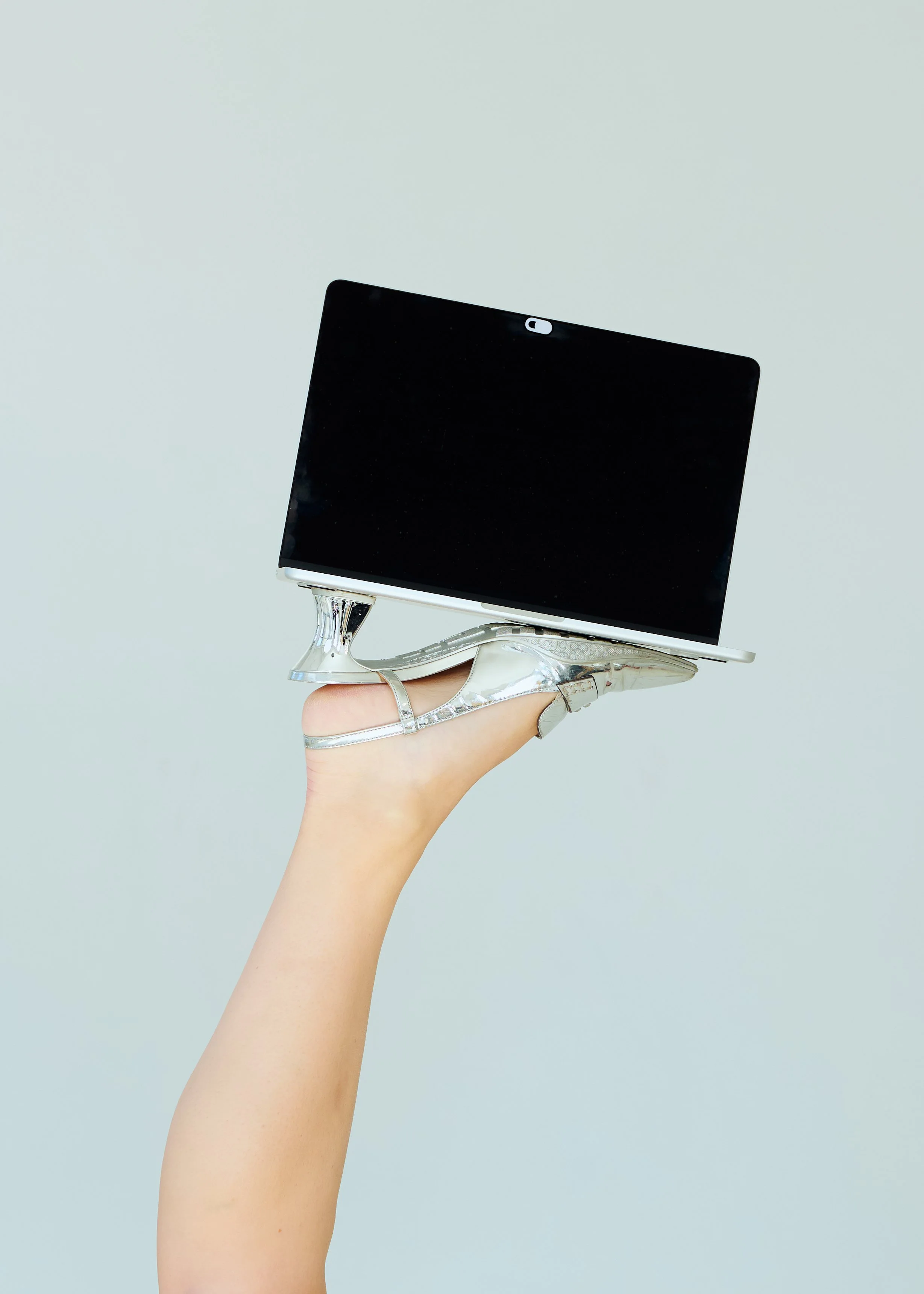 A woman's hand holding a laptop with a stylish silver high-heeled shoe as the sole against a plain, light-colored background.