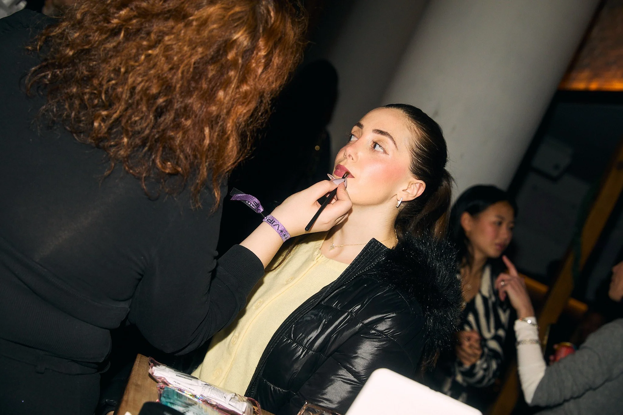Makeup artist applying lipstick to a young woman at a beauty event.
