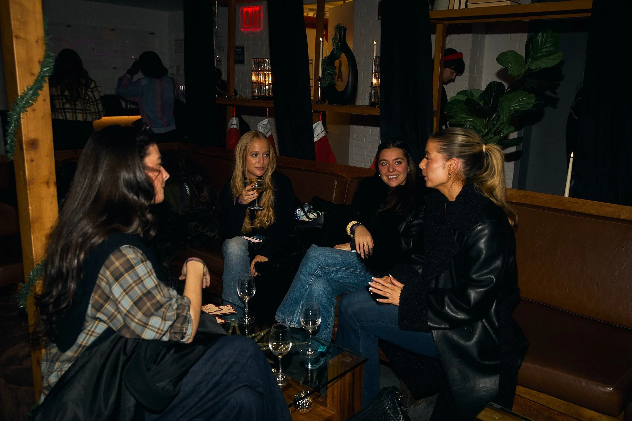 Five women sitting on a brown leather couch in a dimly lit bar or restaurant, engaged in conversation, with glasses of wine and champagne on a table in front of them. Decorations include holiday stockings and candles, suggesting a festive atmosphere.