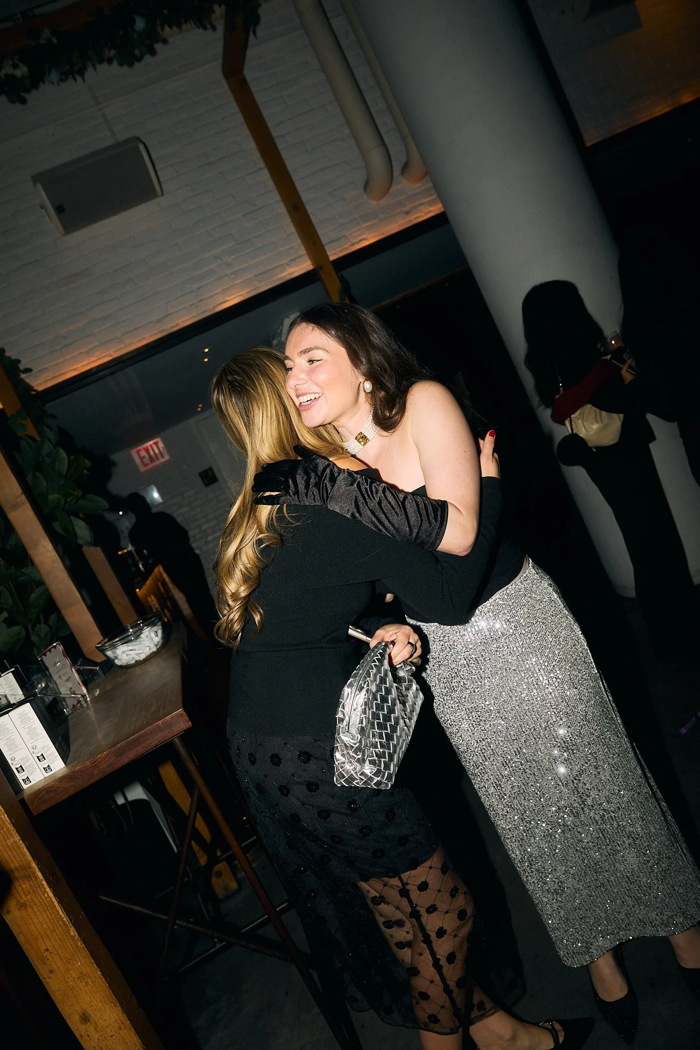 Two women embracing and smiling in a dimly lit indoor setting, possibly a party or event, with one woman in a black dress and the other in a sparkly silver skirt.