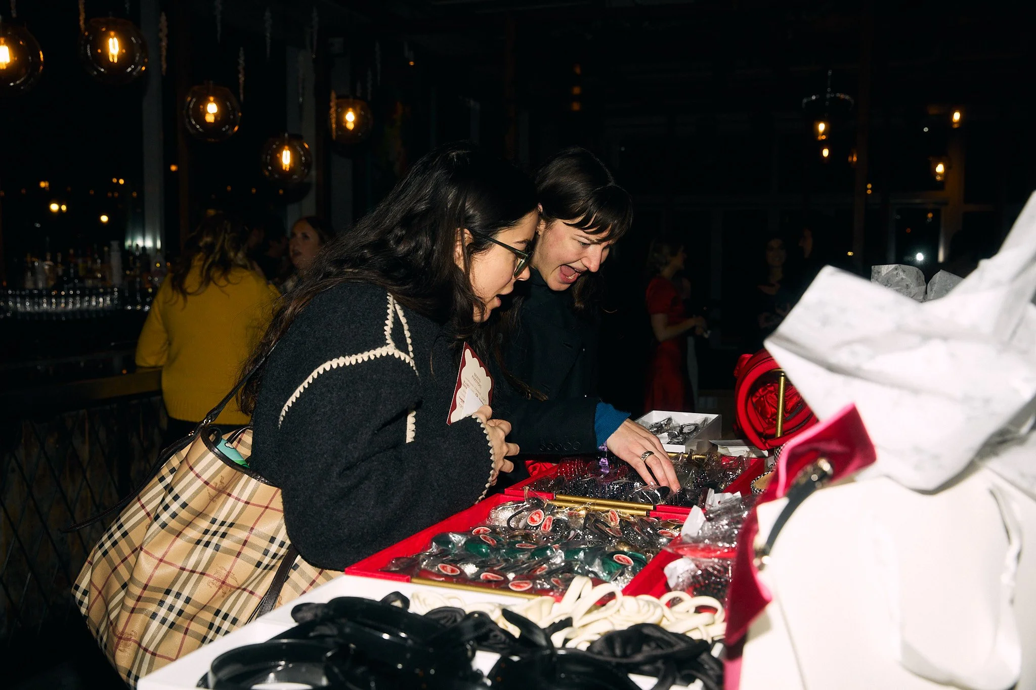 Two women shopping for jewelry at a market stall, looking at assorted jewelry pieces on display.