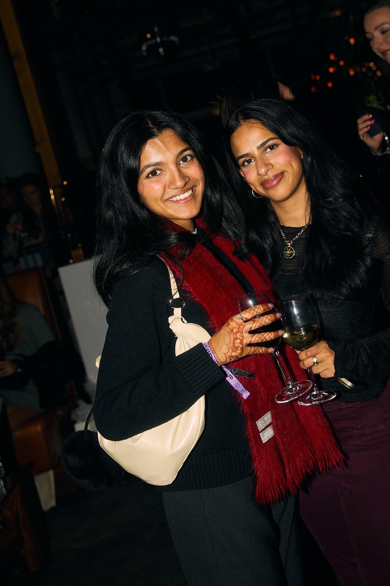 Two women holding glasses of white wine at a social gathering or party, smiling at the camera, with dark indoor background.