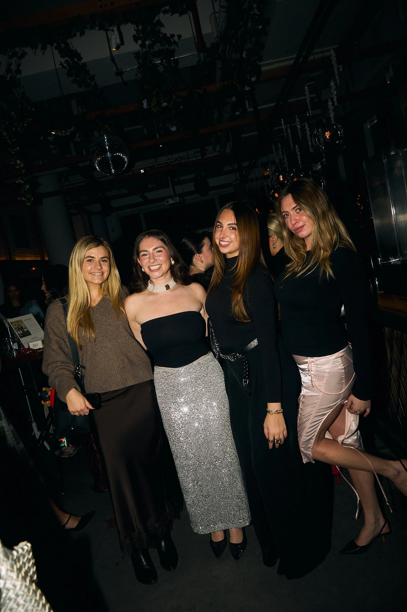 Four women standing together at a night event, smiling at the camera, dressed stylishly with a dark background and dim lighting.