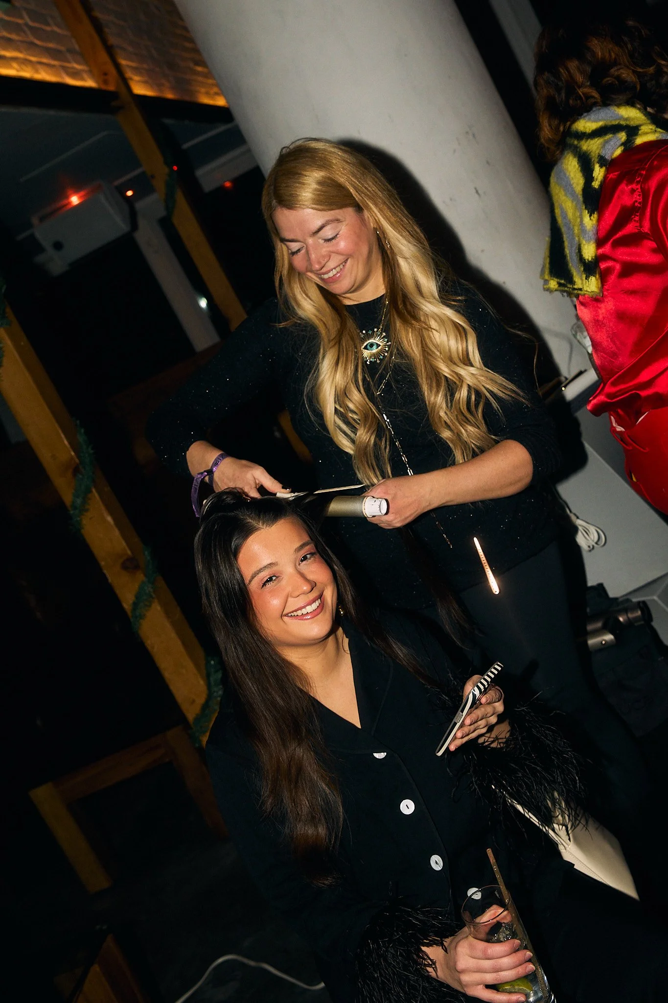 Two women at a party, one has long blonde hair and is styling the other woman with a hair curling iron, who is holding a drink and smiling at the camera.