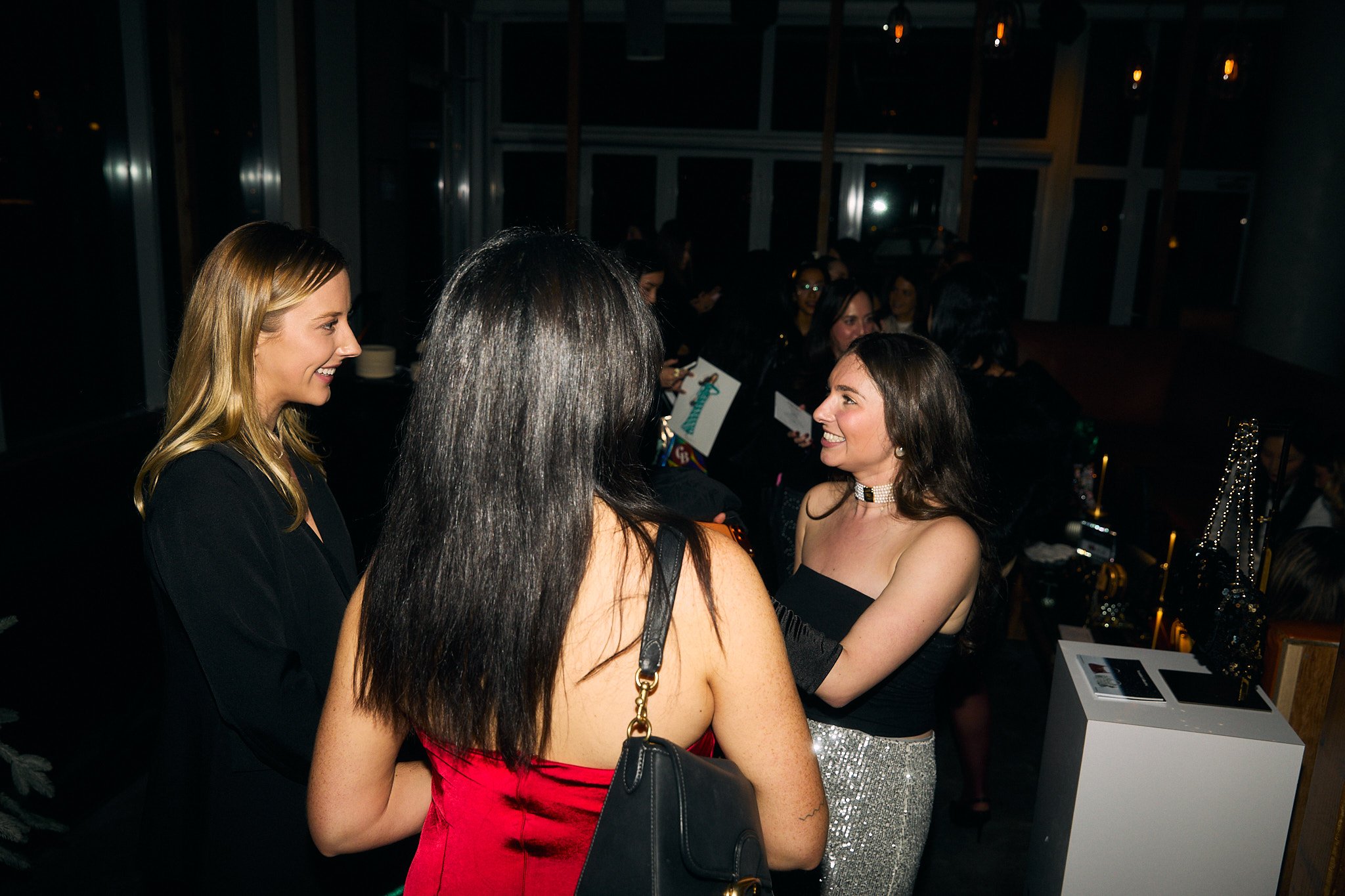 Three women in conversation at a social event, two women facing each other with smiling faces and the third woman with her back to the camera. One woman wears a black blazer, another a red dress with a black purse, and the third a black strapless top