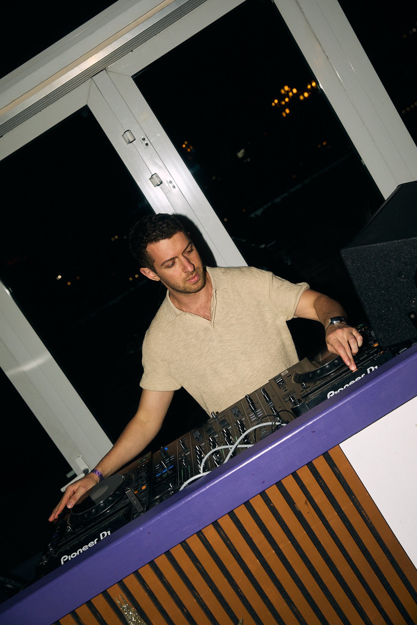 A young man DJing at night, using Pioneer DJ equipment indoors with city lights visible through windows behind him.