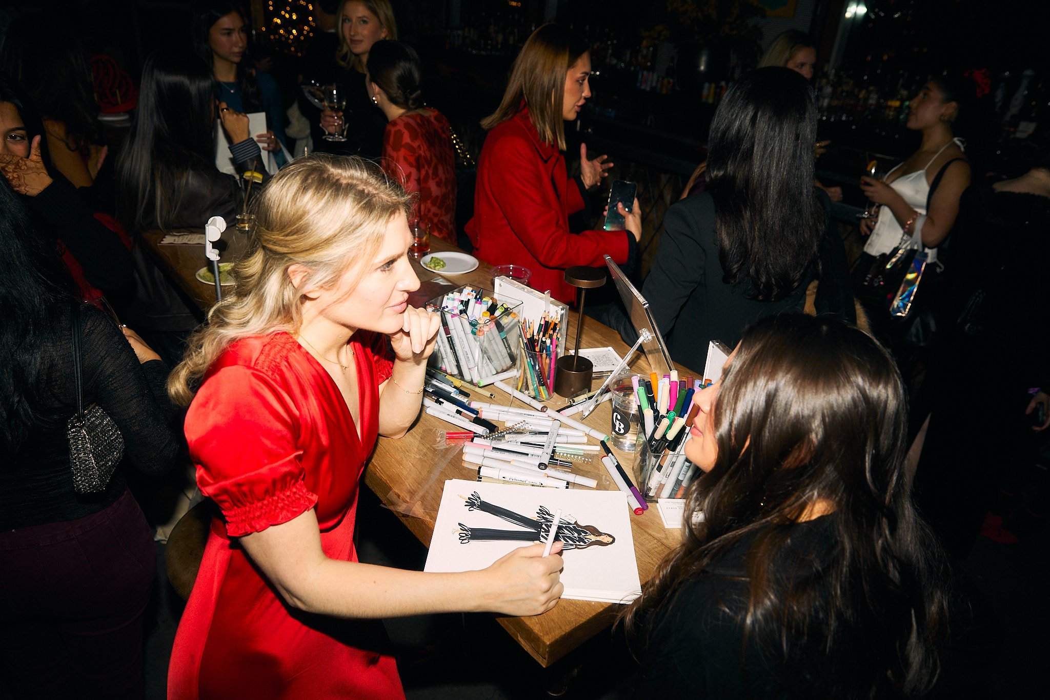 A woman in a red dress sitting at a table with art supplies, sketching a fashion illustration, surrounded by other women at a social event.