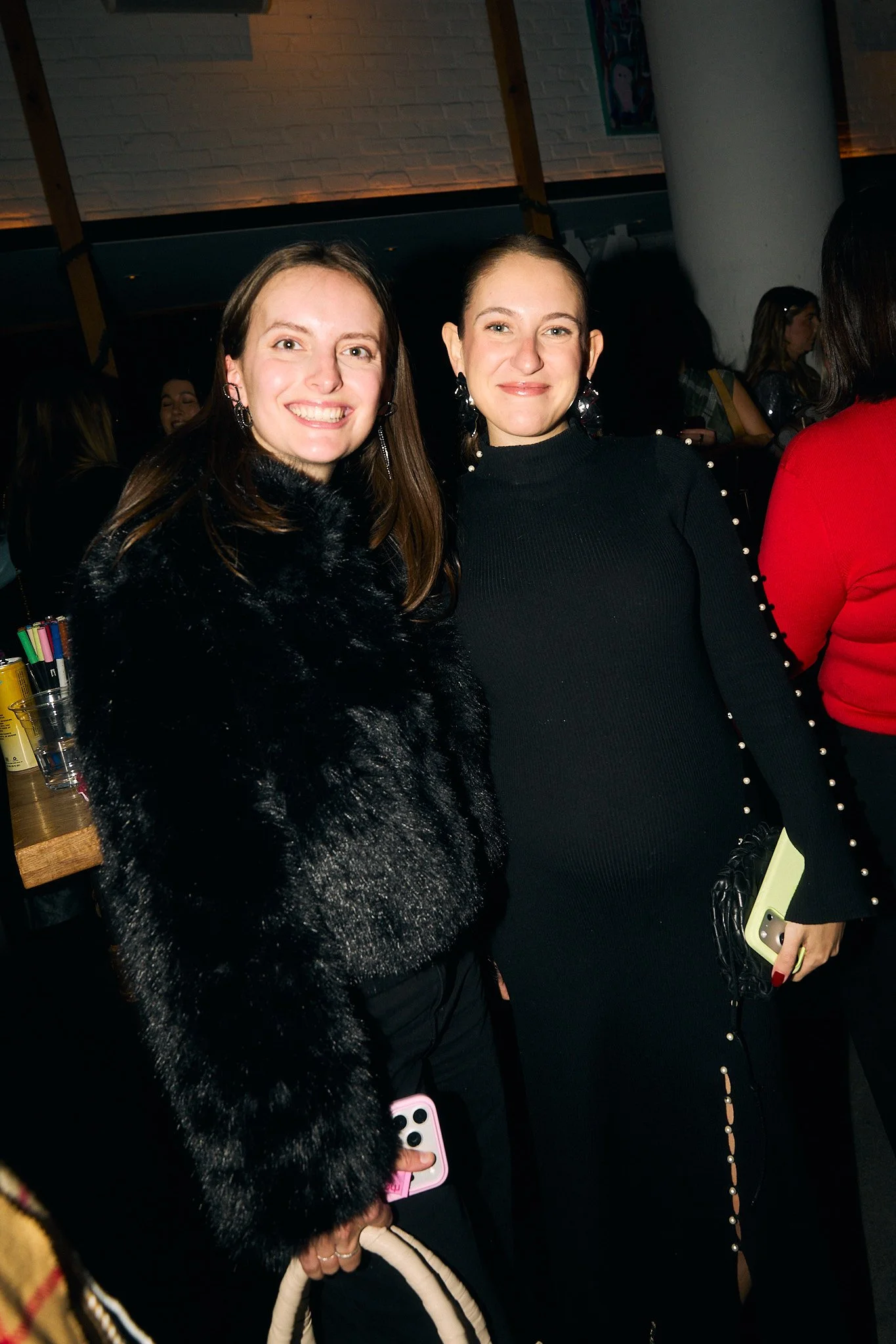 Two women standing close together in a social setting, smiling at the camera, with dim lighting and other people in the background.
