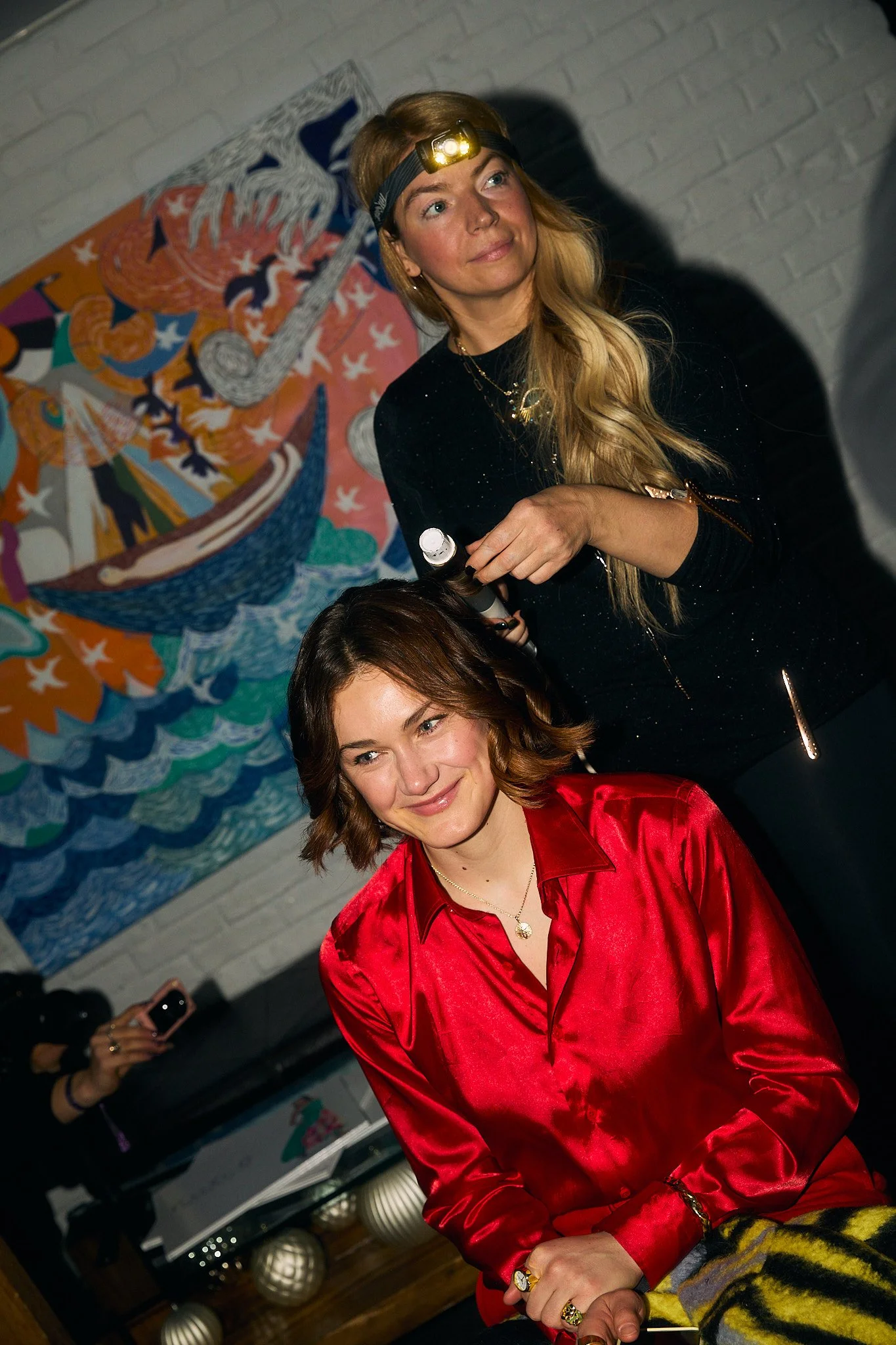 A woman with shoulder-length brown hair wearing a red satin shirt sits smiling while a hairstylist with long blonde hair, wearing a black long-sleeve top, uses a curling iron on her hair. The hairstylist has a headlamp on her forehead. In the backgro