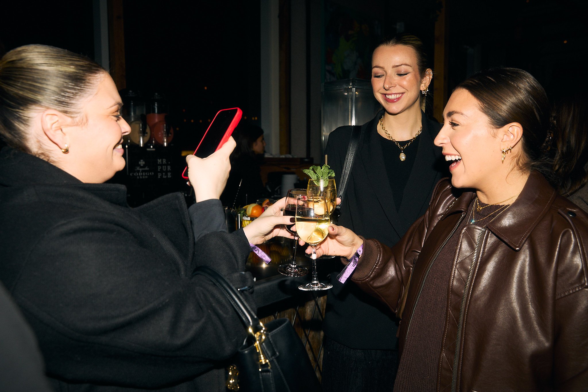 Three women celebrating with glasses of white wine in a bar, smiling and taking a picture together.