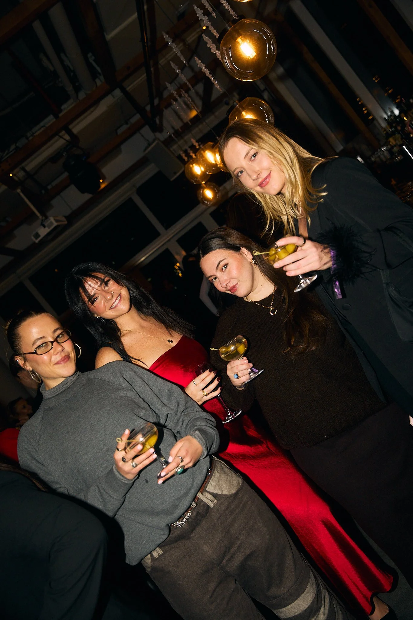 Group of four women at a party or bar, holding glasses of drink, smiling, with warm lighting and modern decor in the background.