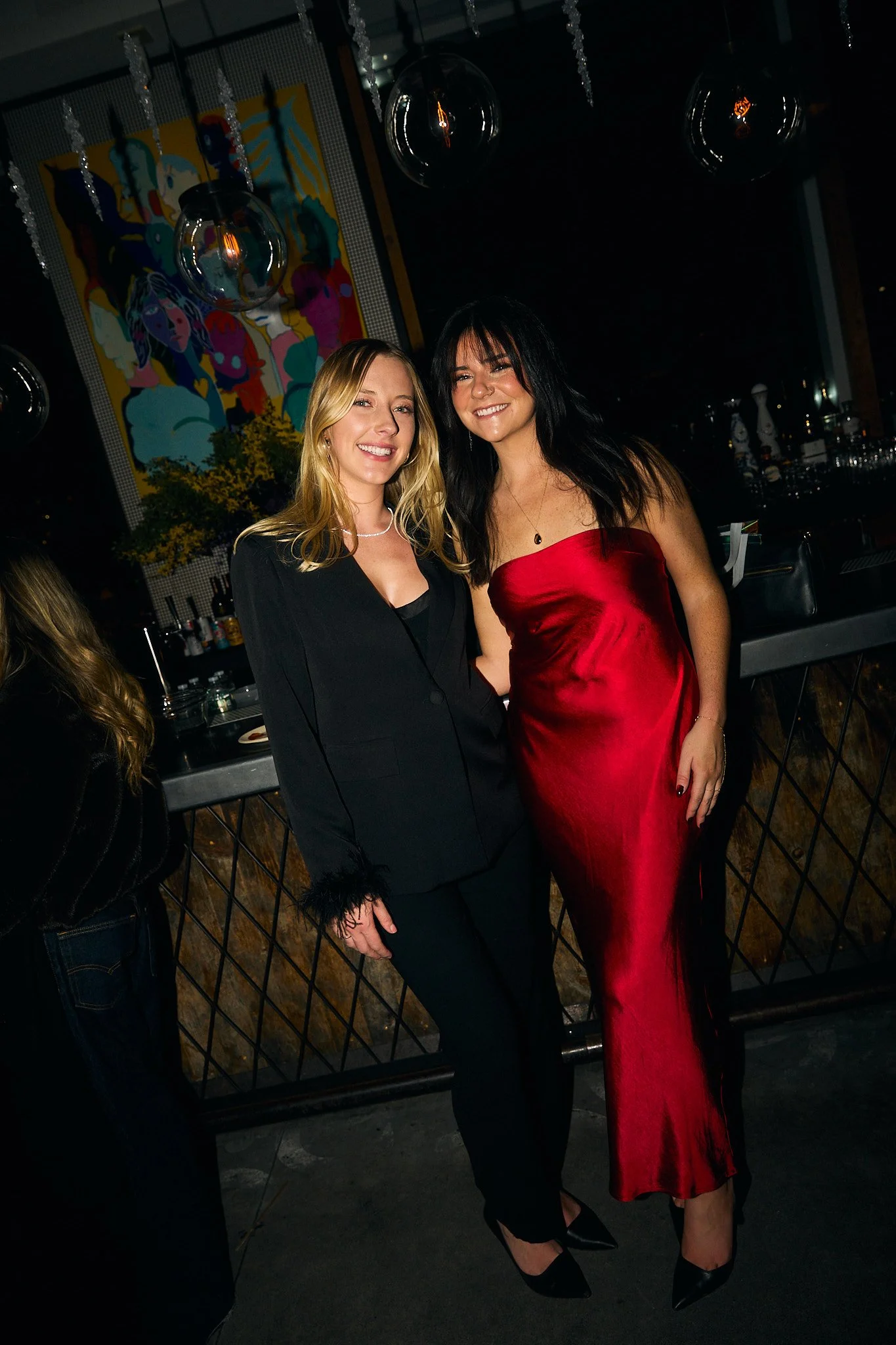 Two women posing together at an indoor event, one in a black suit and the other in a red satin dress.