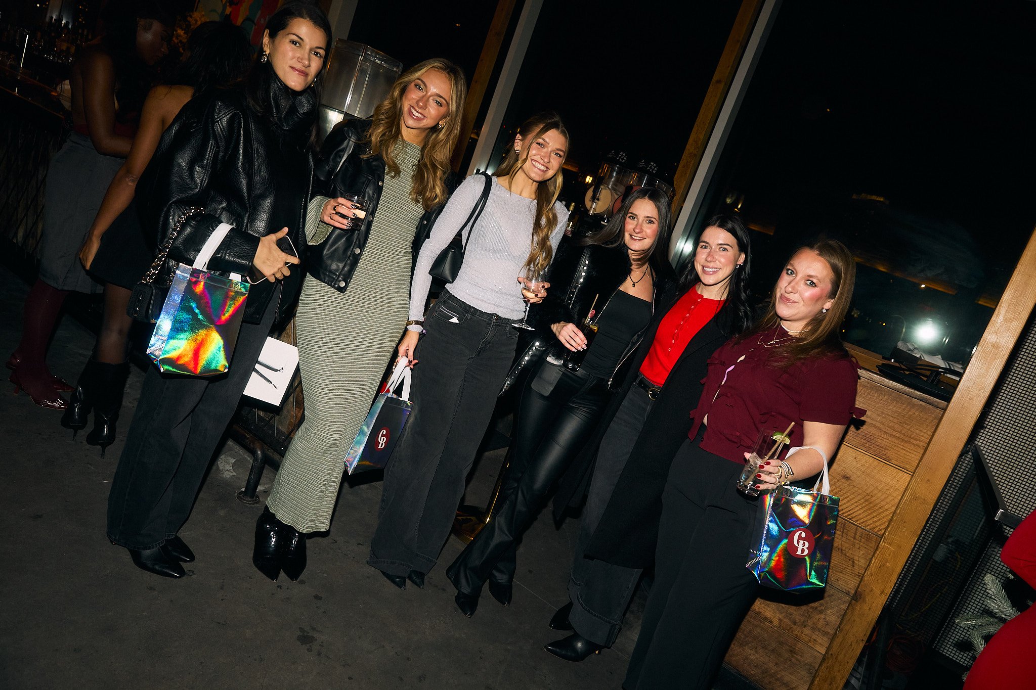 Group of six women at a bar or party, holding drinks and posing for a photo, dressed in stylish outfits, in a dimly lit indoor setting.
