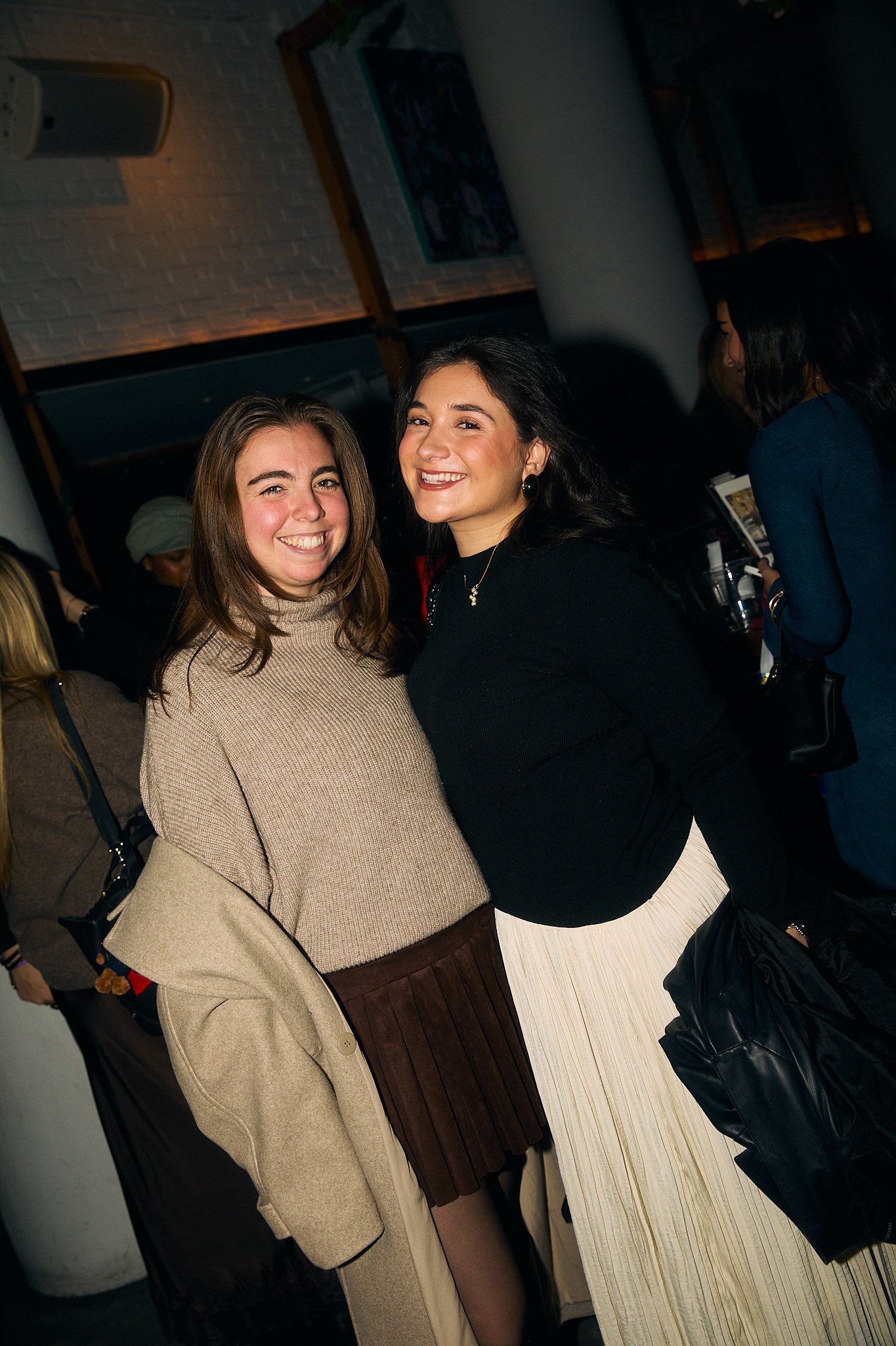 Two women smiling and posing together at a social gathering indoors.