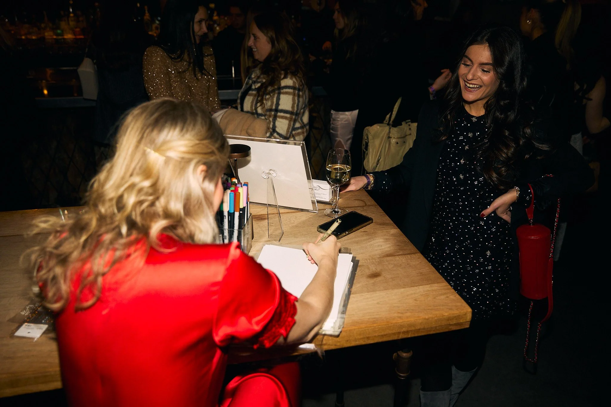 Two women are sitting at a wooden table in a crowded indoor venue. One woman, with blonde hair and wearing a red satin garment, is writing or signing on a notebook. The other woman, with dark hair and dressed in dark clothing, is smiling and holding 