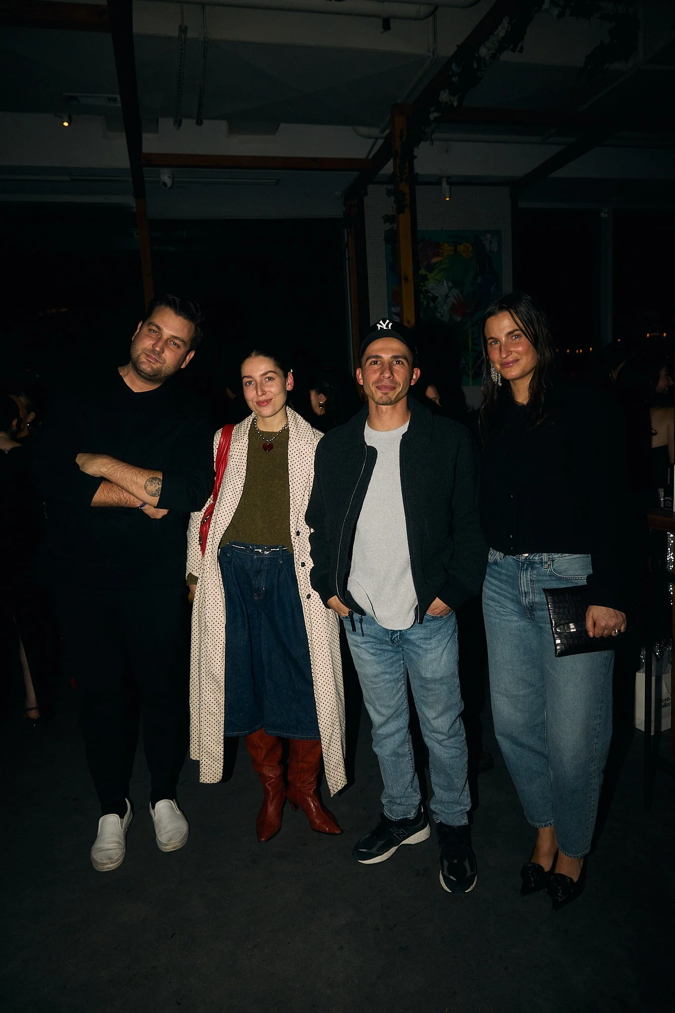 Four people standing closely together at an indoor event, smiling at the camera. The background is dark with some artwork and structural elements visible.