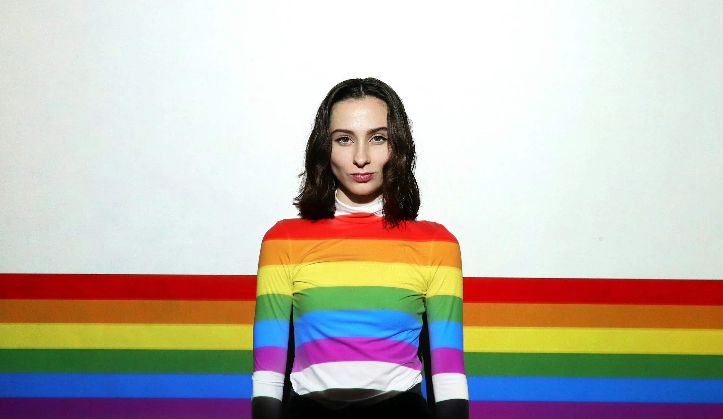 A woman with shoulder-length dark hair wearing a rainbow-striped shirt with red, orange, yellow, green, blue, purple, and white horizontal stripes, standing against a white background with a rainbow stripe pattern on the wall behind her.