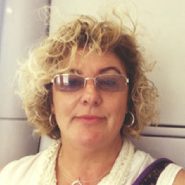 A woman with curly blonde hair wearing sunglasses, hoop earrings, a necklace, and a white top, smiling slightly, sitting indoors.