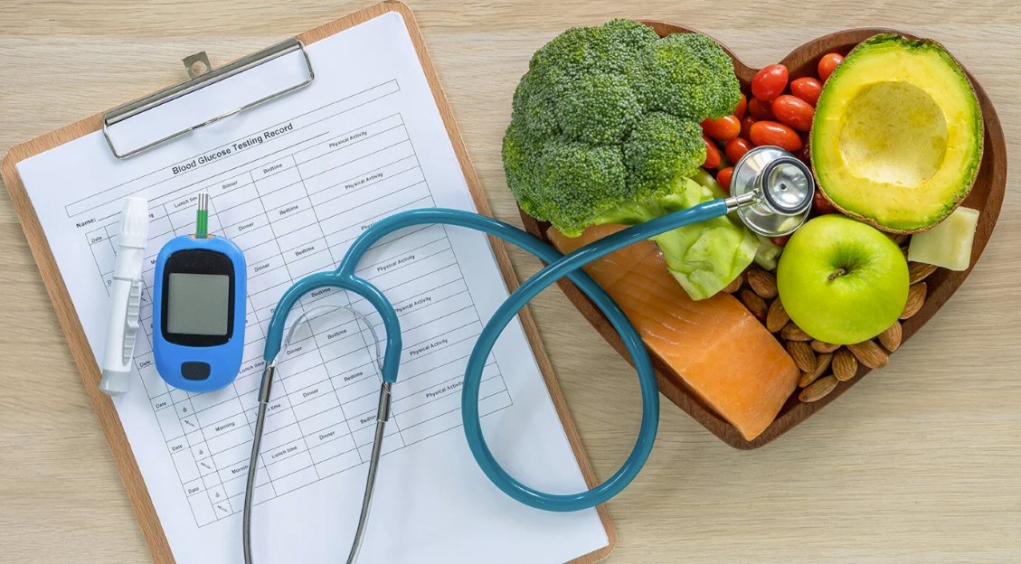 A blood glucose testing record sheet, a glucometer, and medical stethoscope placed on a clipboard, with a heart-shaped wooden bowl filled with healthy foods including broccoli, cherry tomatoes, an avocado, apple, salmon, nuts, and some cheese.